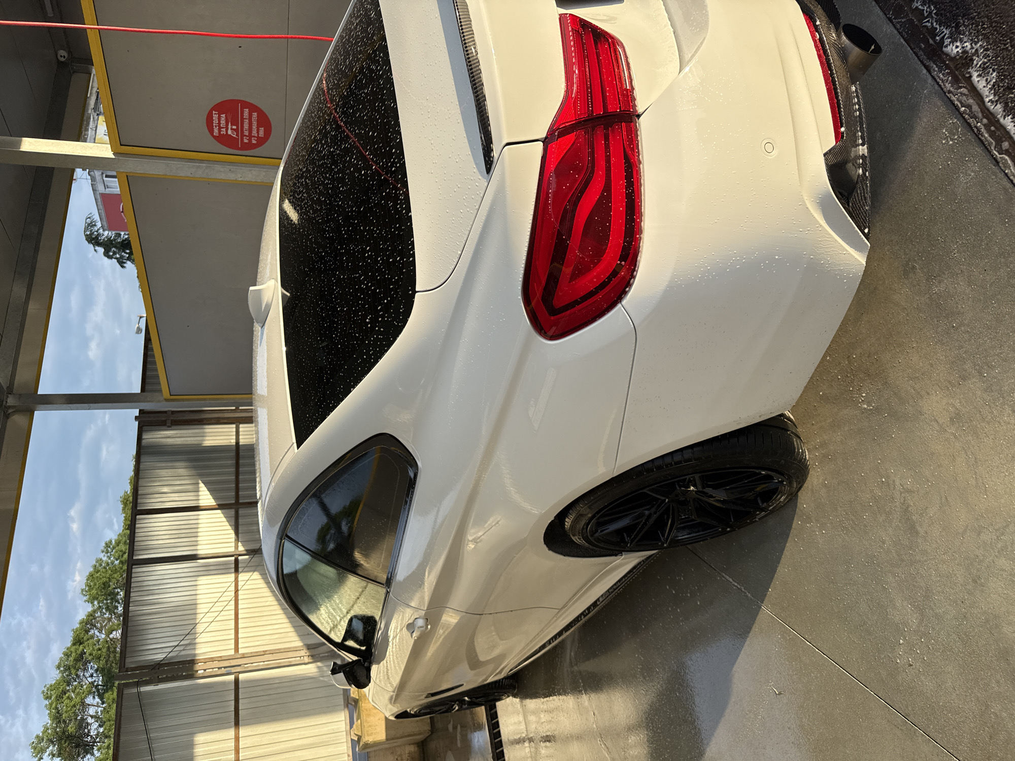 The image displays a white, sporty coupe or sedan undergoing a wash at an outdoor car wash bay in Varna, Bulgaria, under bright daylight. The car's rear-right section is prominently featured, appearing wet with water droplets and some soap residue, suggesting it is either being washed or has just been rinsed. Notable details of the car include a distinct red LED taillight, a black multi-spoke wheel, a dark exhaust tip, and what appears to be a carbon fiber diffuser or trim around the exhaust. A parking sensor is also visible on the white bumper.
The car wash facility includes a wet concrete floor and a structure with a yellow beam from which a red hose hangs. A red circular sign attached to the overhead structure provides instructions and options in Bulgarian. The text reads: "ПИСТОЛЕТ ЗА ПЯНА" (FOAM GUN), "№1 АКТИВНА ПЯНА" (№1 ACTIVE FOAM), and "№2 ДИАМАНТЕНА ПЯНА" (№2 DIAMOND FOAM).
In the background, a corrugated metal fence or wall is visible, with a clear blue sky and green trees beyond it. The presence of distinct shadows on the ground and car indicates direct sunlight, suggesting a morning or late afternoon setting. No individuals are present in the scene.