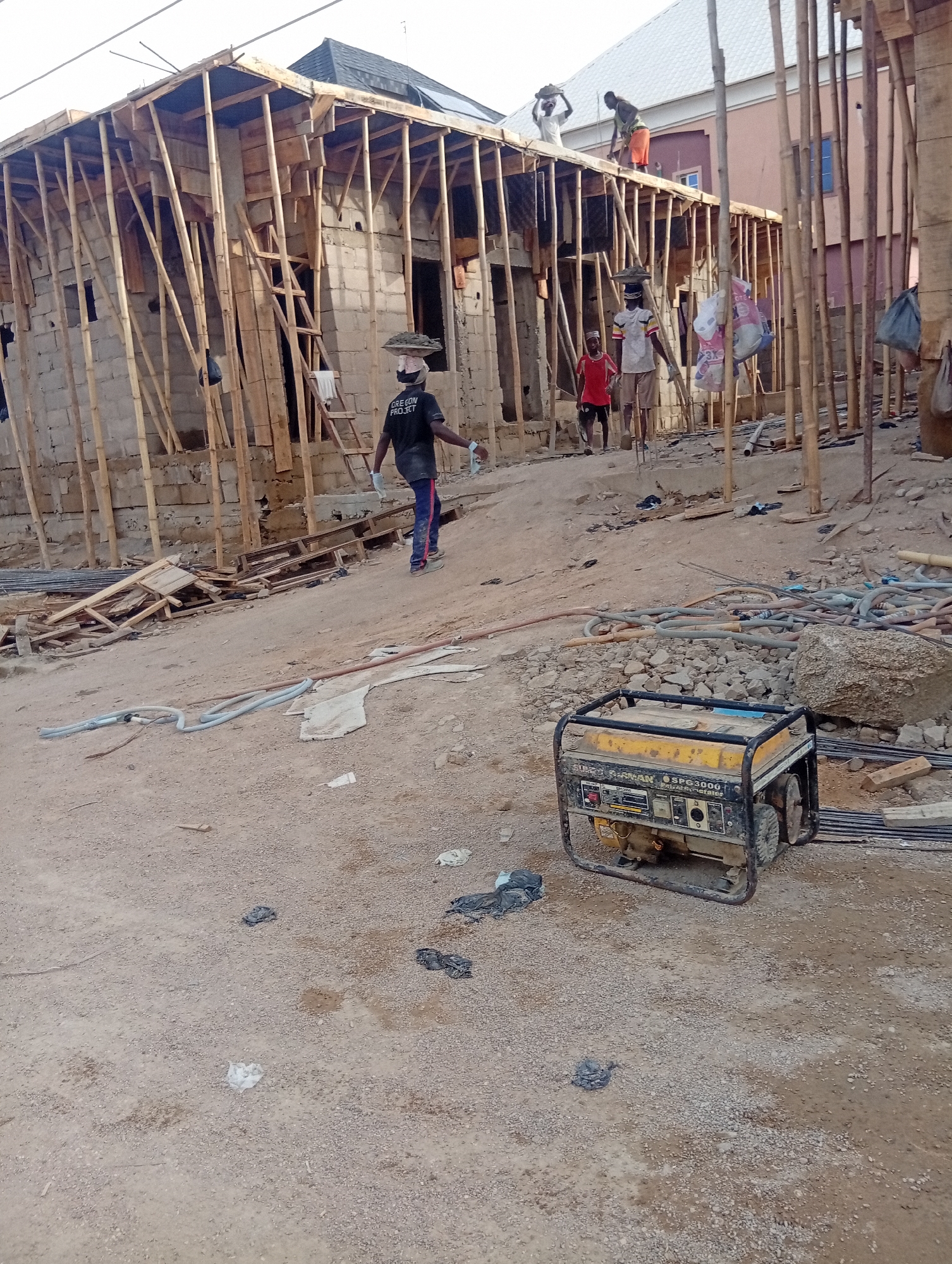 The image shows a construction site in Aviation Quaters, Nigeria. A partially constructed building is visible, supported by wooden scaffolding. Several people are present, including construction workers, one of whom is carrying material on his head. A portable generator sits in the foreground amidst construction debris, including metal rods, hoses, and scattered plastic bags. The ground is dirt and gravel.