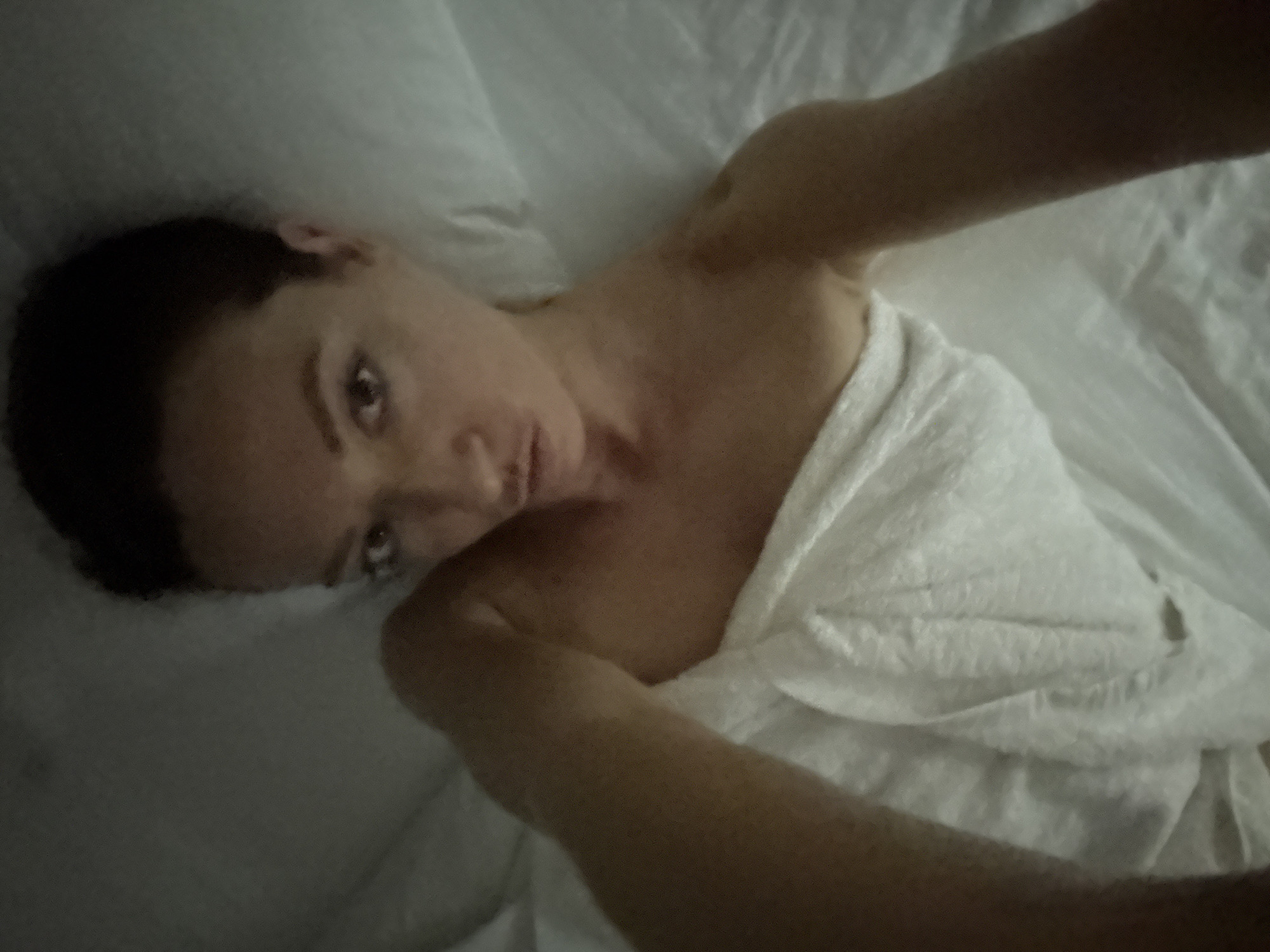 An individual, likely female, with dark hair and fair skin, is depicted in a close-up, high-angle self-portrait. She is positioned supine or semi-supine on a bed with her head resting on a white pillow, looking directly into the camera. Her lips are pursed. She is wrapped in a white fabric, possibly a sheet or towel, which covers her body from the upper chest downwards, leaving her shoulders and arms bare. Her left arm (viewer's right) is extended towards the camera, while her right arm (viewer's left) is bent with the hand mostly out of frame. The background consists of crumpled white bedding, including sheets and pillows. The lighting is dim and even, suggesting an indoor environment with low illumination, consistent with a bedroom. The scene is described as taking place in the city of Bản Đêu, Vietnam.