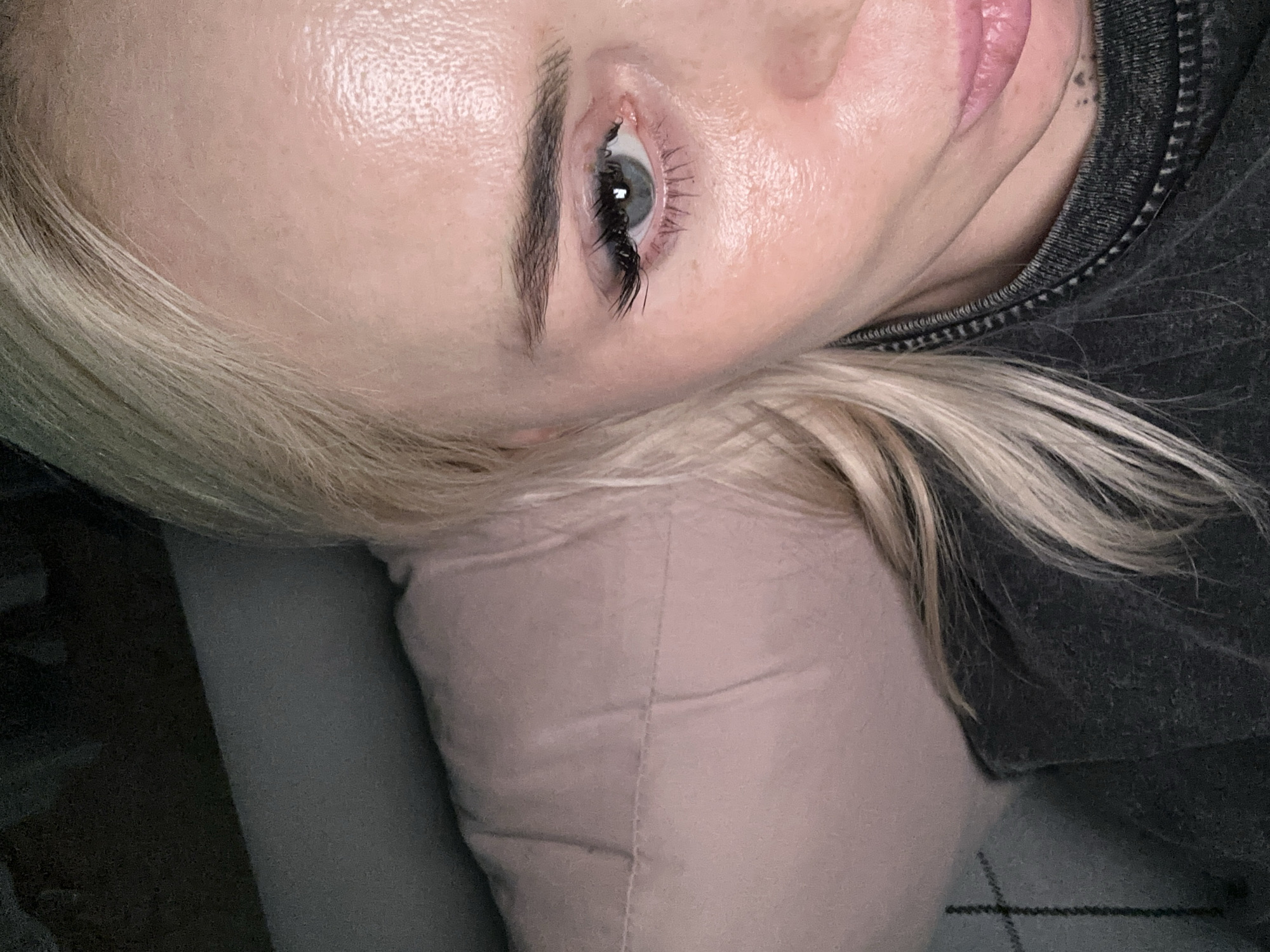 This close-up, angled portrait captures the face of a person, likely female, from an overhead perspective while they are lying down. The dominant features are a striking blue or green eye, the forehead, part of the nose, and lips.
The subject has fair, dewy skin, which shows a hydrated and slightly reflective texture, particularly on the forehead and around the eye. A dark, well-defined eyebrow arches above the open eye. Long, dark eyelashes, possibly enhanced with mascara or extensions, frame the eye, and a distinct reflection is visible in the pupil. Below the eye, the upper part of the nose is visible, leading to slightly parted, light pink lips on the right side of the frame. A faint tattoo, consisting of small dark dots, is visible on the neck/jaw area beneath the earlobe.
Blonde hair cascades around the head, visible along the bottom and left edges of the frame. The person is wearing a dark, textured garment, possibly a hoodie or jacket, with a zipper detail noticeable around the collar. Their head rests on a light pink or beige pillow, which exhibits some creasing. The background is softly blurred, showing muted dark and grey tones, suggesting an indoor setting like a bed or couch. The overall impression is one of calm and tranquility. No text is visible in the image.