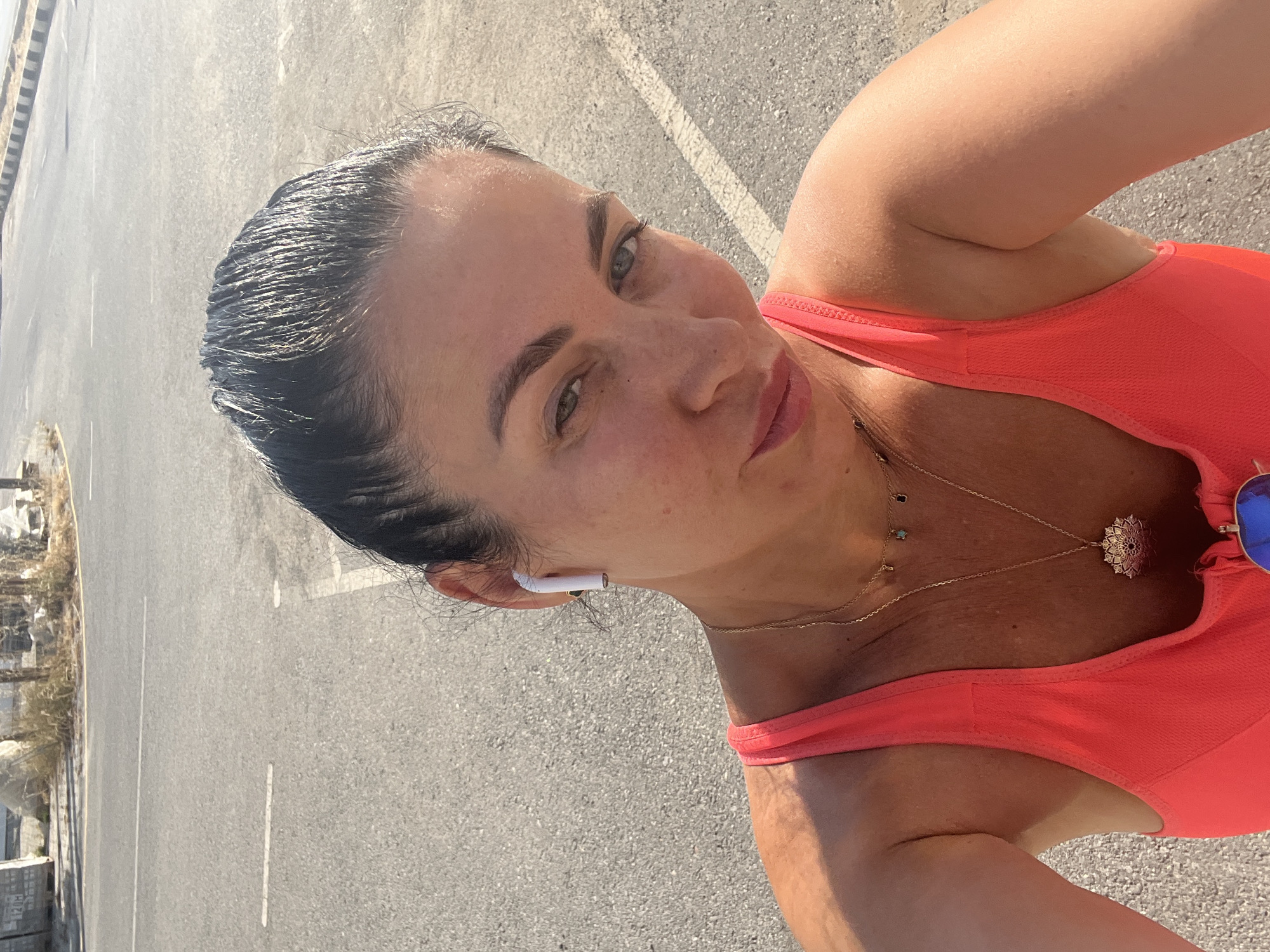 A woman is captured in a close-up, upward-angled selfie on an asphalt road in Trikomo, Northern Cyprus. Her dark hair is pulled back, appearing slightly damp and reflecting sunlight. She has light-colored eyes, visible eyebrows, and is wearing light-toned lipstick. A white wireless earbud is visible in her right ear. She wears a bright coral-orange sports bra or tank top. Adorning her neck are multiple gold-colored necklaces: a shorter one with small, dark and turquoise-colored charms, and a longer one featuring a large, ornate, circular pendant. Blue mirrored sunglasses are clipped to the front of her top, partially visible at the bottom. The background consists of a grey asphalt road with white painted lines. In the far distance, dry vegetation and some indistinct light-colored structures or debris are visible under bright daylight conditions.