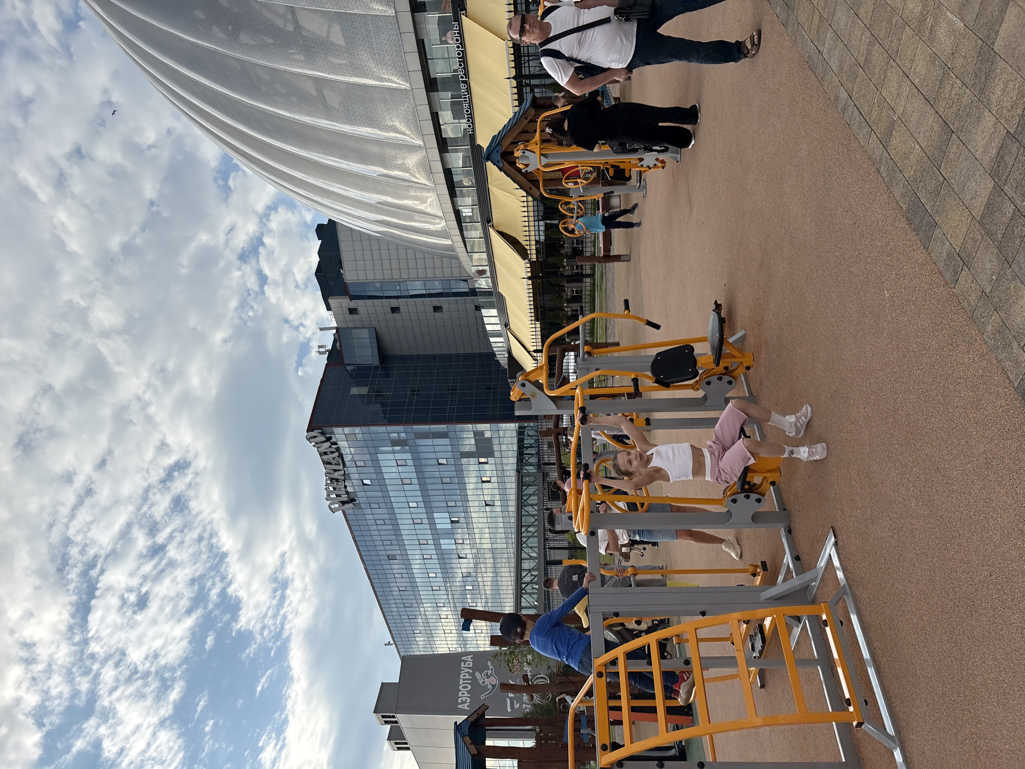 This image captures an outdoor recreational area in Saint Petersburg, Russia, featuring people utilizing exercise equipment amidst modern architecture under a bright, cloudy sky.
In the foreground, several individuals are present, with a young girl in a white crop top and pink shorts lying on a yellow and grey outdoor exercise machine, and a boy in a blue long-sleeved shirt at another. Other people, including an adult male in a white t-shirt and sunglasses, are standing or walking nearby, observing the activities. The ground is covered with a soft, reddish-brown, textured surface around the exercise machines, transitioning to a patterned paved area on the right.
The background is dominated by large, contemporary buildings. A tall, dark-colored building with a facade of numerous reflective windows stands prominently, reflecting the sky. Part of a large, translucent white architectural structure, possibly a canopy or dome, is visible on the left. Below this structure, an awning bears the Cyrillic text "настоящие рестораны" (real restaurants). To the left, a smaller building features the Cyrillic text "АЭРОТРУБА" (aerotube or wind tunnel). The main dark building also displays the text "ГАЗПРОМ" near its top.
The scene takes place during the daytime under a bright sky with scattered white clouds, suggesting good weather for outdoor activities. The overall setting is a vibrant urban environment where recreation, dining, and modern architecture converge.