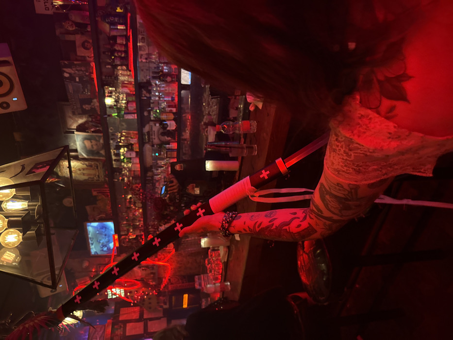 A blurry image captures a person in a dimly lit bar, holding a sword-like object decorated with crosses. The individual has extensive tattoos on their arm, and is wearing a lacy white top. The bar backdrop features numerous bottles behind the counter, ambient lighting, and a small screen displaying content. A decorative light fixture hangs on the left, adding to the overall low-light atmosphere.