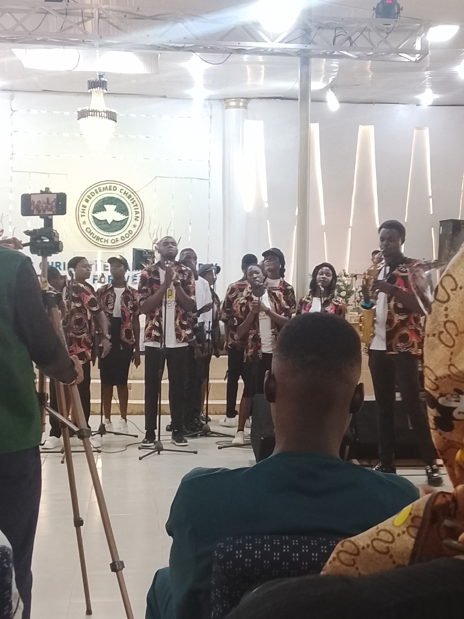 A medium shot captures a scene indoors, likely a church, as indicated by the "The Redeemed Christian Church of God" logo prominently displayed on a white wall in the background. An ensemble of approximately ten people stands on a stage, dressed in patterned shirts over white t-shirts, and dark pants or skirts. They appear to be singing or performing, with microphones positioned in front of them. One individual on the right holds a saxophone.
In the foreground, the backs of several audience members are visible, including a man with a short haircut wearing a blue-green shirt, and another person whose shoulder and bag, adorned with a pattern, are partially in the frame. A video recording setup, consisting of a phone or small camera mounted on a tripod, is visible on the left, suggesting the performance is being recorded.
The environment is bright and appears well-lit with an overhead chandelier and stage lighting. Vertical white panels decorate the wall behind the stage.