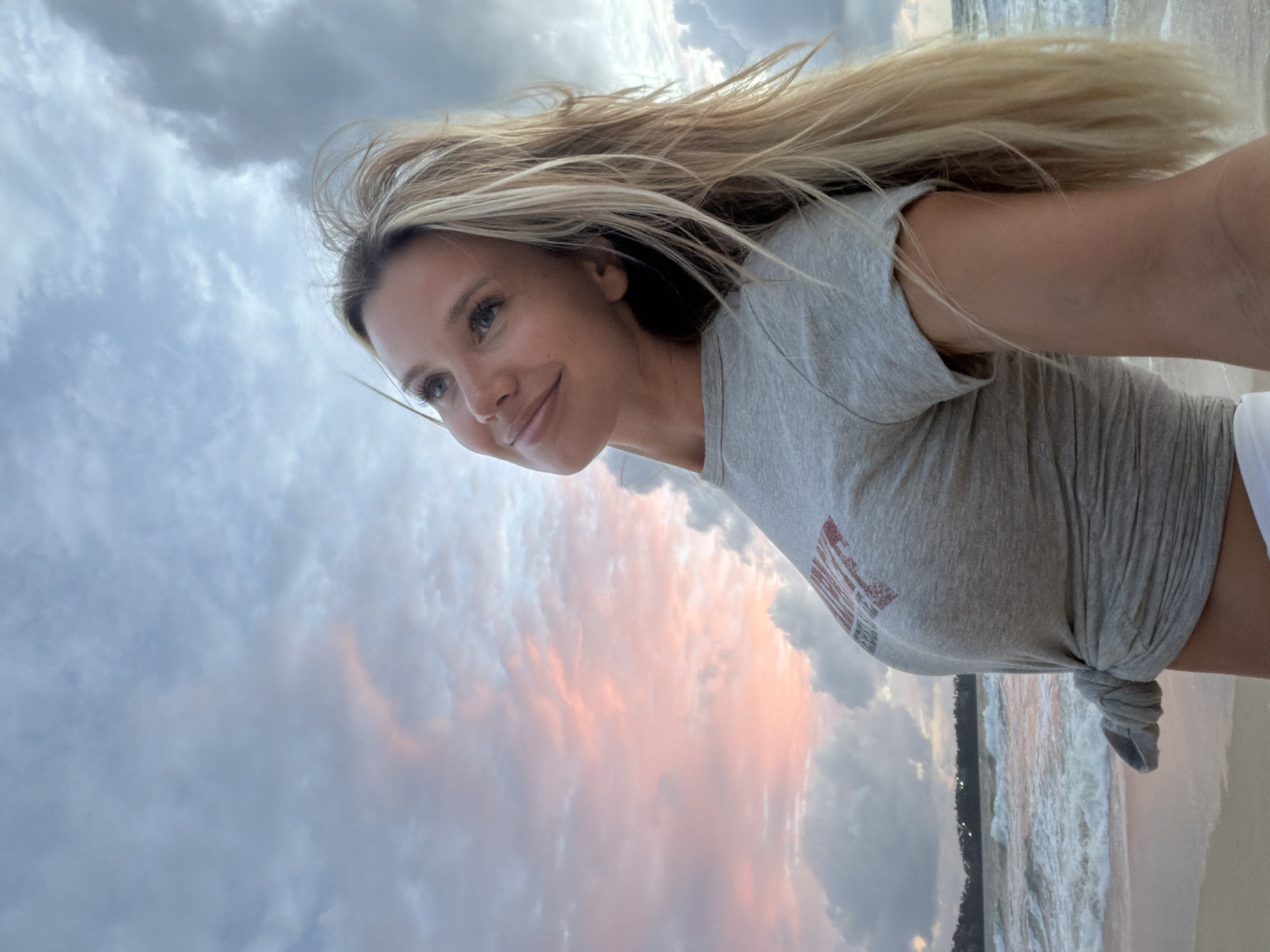 A young Caucasian woman with long blonde hair and blue eyes is standing on a beach in Choeng Thale, Thailand, looking up and to her left with a slight smile. She is wearing a grey t-shirt tied at her waist and white shorts. The ocean with waves is behind her, and the sky is filled with dramatic clouds, with hints of pink and orange from a sunset. The sand is visible in the foreground.