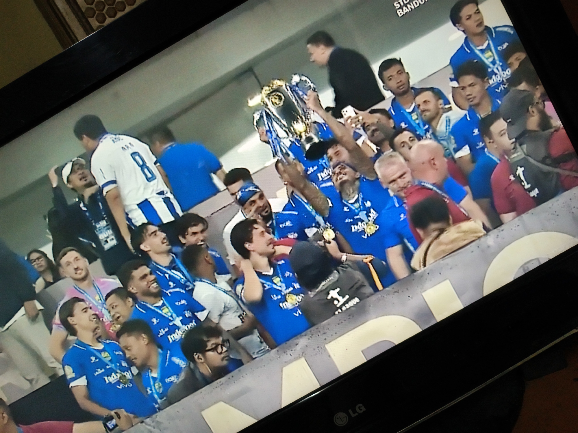 Captured on an LG television, the image depicts a celebration, presumably related to a soccer team winning a trophy, based on the presence of a large trophy being hoisted aloft and a group of people, primarily wearing blue jerseys, jubilantly surrounding it. Most of the individuals are of Asian ethnicity. They are standing closely together, with a few people of Caucasian descent also in the mix. The setting appears to be within or near a stadium, perhaps during a ceremony or immediately following a game. The image's top right corner bears some text ending in "BANDUN", suggestive of Bandung, Indonesia. Pacet is not visible in the image, nor can its presence be deduced based on the photo.