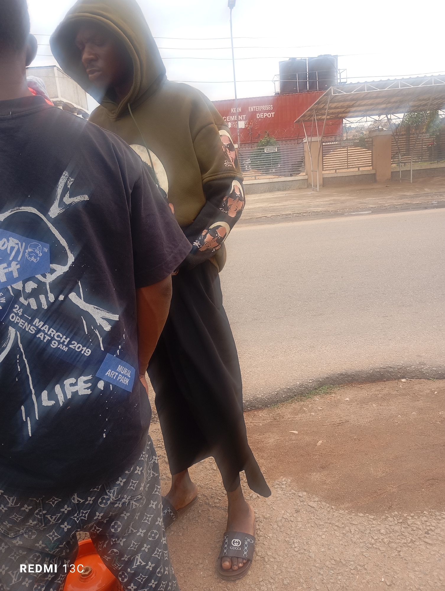 Two individuals stand on a dirt and gravel roadside in Jos, Nigeria. The person in the foreground, facing away from the camera, wears a black t-shirt with white graphic designs and text that reads "LIFE" and "MURAL ART PARK" and "24 - MARCH 2019 OPENS AT 9 AM", and patterned trousers with a repeating Louis Vuitton logo. The second individual, to the right and partially obscured, wears a green hooded sweatshirt with graphics on the sleeves and black pants, and black Gucci slides. In the background, a large red shipping container serves as a storefront for "KE.IN ENTERPRISES CEMENT DEPOT". A covered awning area is visible in front of the depot. The sky is overcast.