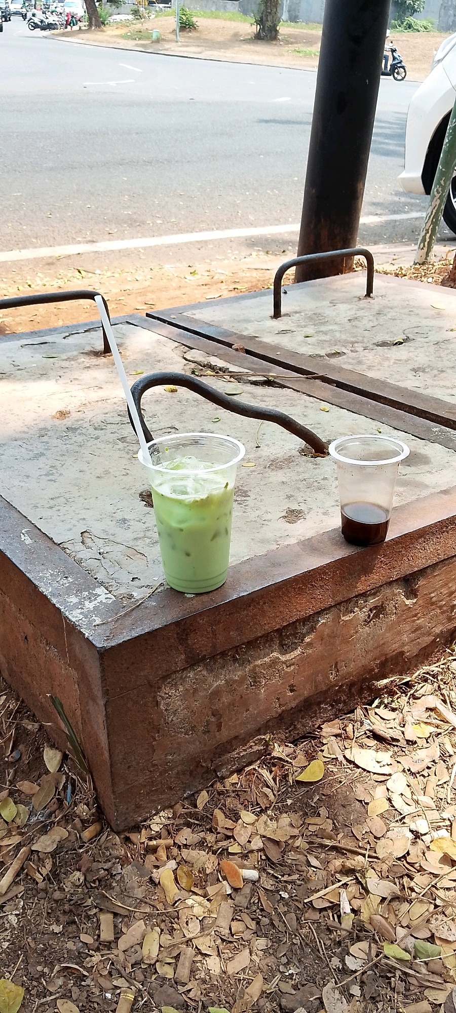 The image depicts two plastic cups of drinks on a concrete surface in an outdoor urban environment, likely near a street in Gunung Putri, Indonesia. The cup on the left contains a light green beverage, possibly matcha or a similar iced drink, with ice cubes visible. A white straw extends from this cup. The cup on the right contains a dark liquid, resembling coffee or iced tea. Both cups are clear plastic and appear to be disposable.
The concrete surface is weathered and shows signs of wear, with metal grates or fixtures embedded in it. The background shows a paved road with traffic, including motorcycles and a white car. Trees and vegetation are visible along the roadside, suggesting a location with greenery. The ground around the concrete structure is covered with fallen leaves and debris, indicating a natural or unkempt area adjacent to the urban setting. The lighting suggests it is daytime, with sunlight casting shadows. There are no people clearly visible in the immediate foreground.