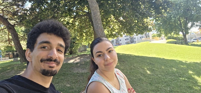 The image captures a selfie of two individuals, a male and a female, in an outdoor park environment under clear, sunny conditions. The male, on the left, has dark curly hair, a mustache, and a goatee, wearing a dark t-shirt, and is smiling towards the camera. The female, on the right, has dark hair pulled back, wears a light-colored patterned sleeveless top, a gold-colored necklace, and red nail polish on her visible right hand, also smiling towards the camera. A large tree trunk stands centrally behind them. The background consists of a sloping grassy area. To the left, a black metal fence partially obscures a children's playground with colorful equipment, beyond which is a multi-story residential building with light-colored siding. To the right, another tree, a wooden picnic table, several parked vehicles (including a white van), and a yellow fire hydrant are visible near a paved surface. The location is identified as Stronach Park in London, Ontario, Canada.