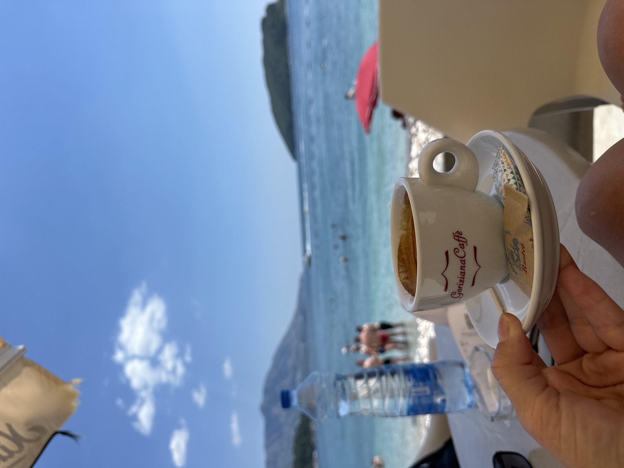 An outdoor cafe table on a beach in Budva, Montenegro, holds a white cup of espresso with the logo "Goriziana Caffè" visible. A hand is holding the saucer of the cup, which also contains a sugar packet. A plastic water bottle is positioned to the left of the cup. In the background, the Adriatic Sea is visible with several people swimming and standing in the shallow water. Mountains rise in the distance, and a small island is situated offshore. The sky is clear blue with a few scattered clouds.