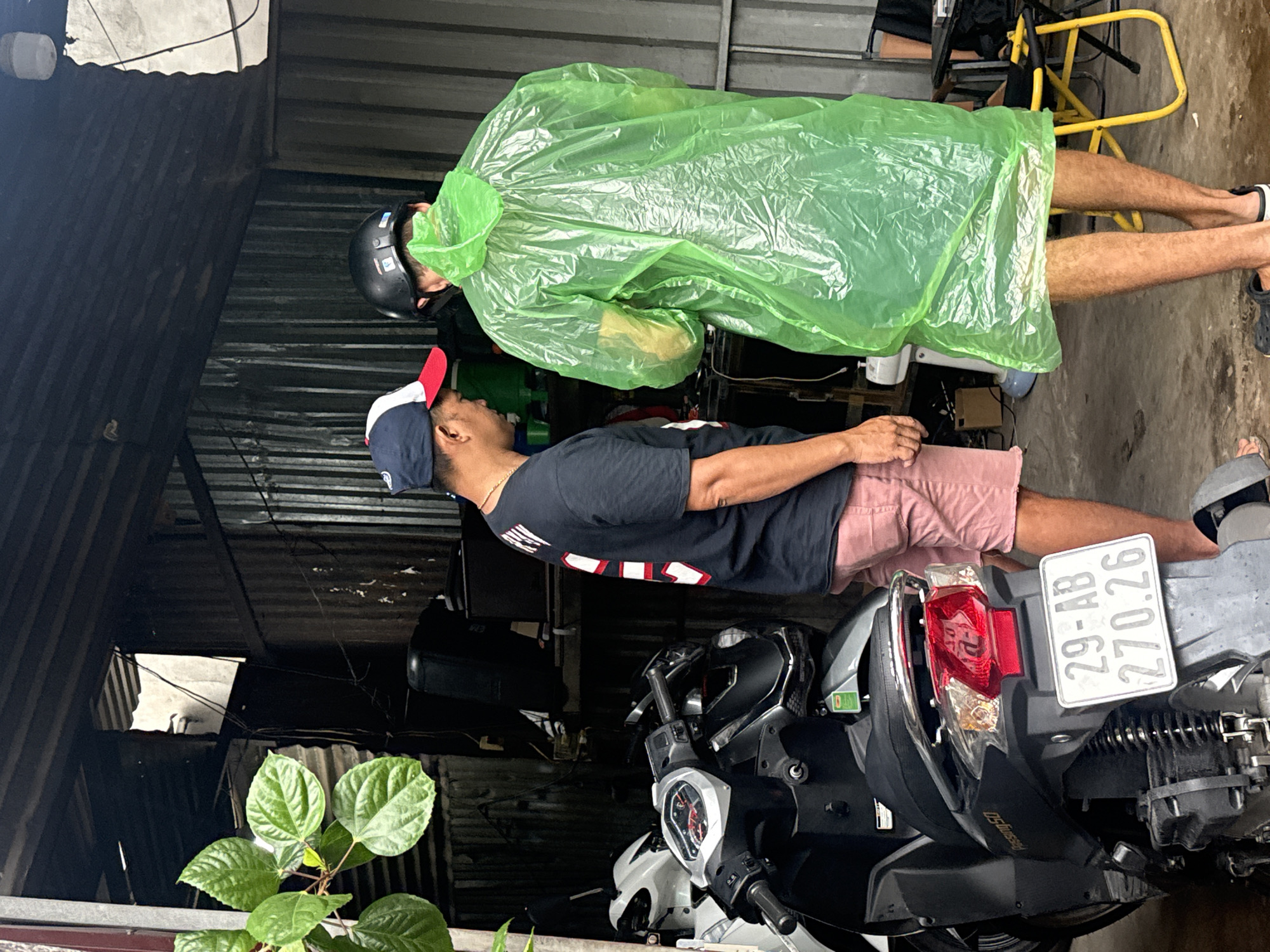 Two men stand under a corrugated metal roof, possibly a carport or workshop, in Nha Trang, Vietnam. The man on the left, wearing a black t-shirt with red and white lettering, a red and white baseball cap, and pink shorts, faces towards the right. The man on the right, seen from behind, wears a black helmet and a bright green plastic poncho. He is facing away from the camera. In the foreground, a dark-colored scooter with a license plate reading "29-AB 270.26" is parked. Behind the men, the environment appears cluttered with various objects, including what looks like electronic equipment and a white fan. Green foliage is visible in the upper left corner. The ground is a concrete or paved surface.