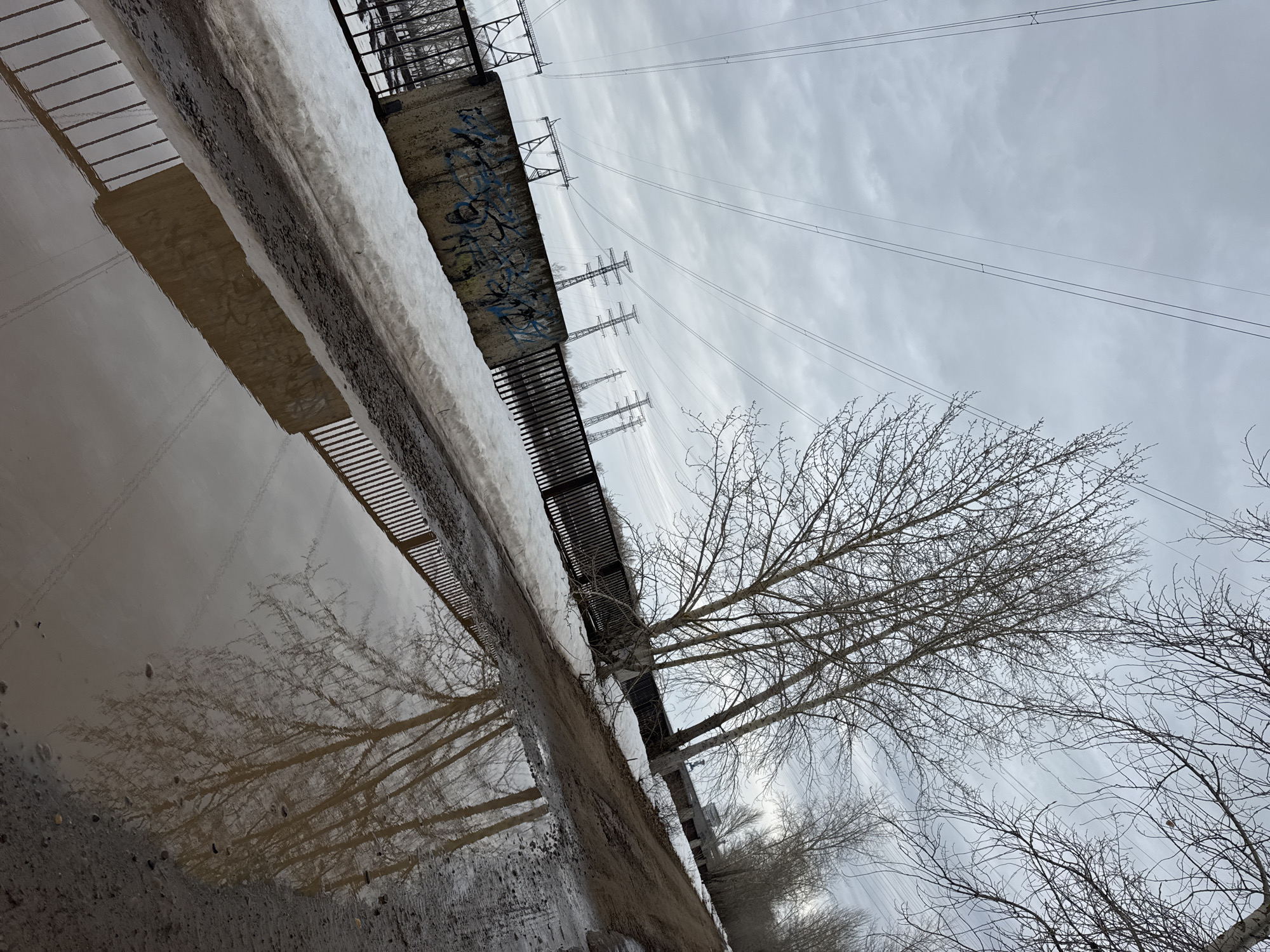 The image captures a damp, overcast scene in what is presumed to be Chaikovsky, Russia. In the foreground, a large puddle reflects elements of the environment, including the bare branches of trees, a metal fence, and a section of a concrete structure possibly graffitied. Patches of snow are visible next to the concrete structure, which appears to be an elevated walkway or platform. In the background, there are power lines and transmission towers against a backdrop of a cloudy sky. The overall impression is that of a cold, possibly post-winter landscape.