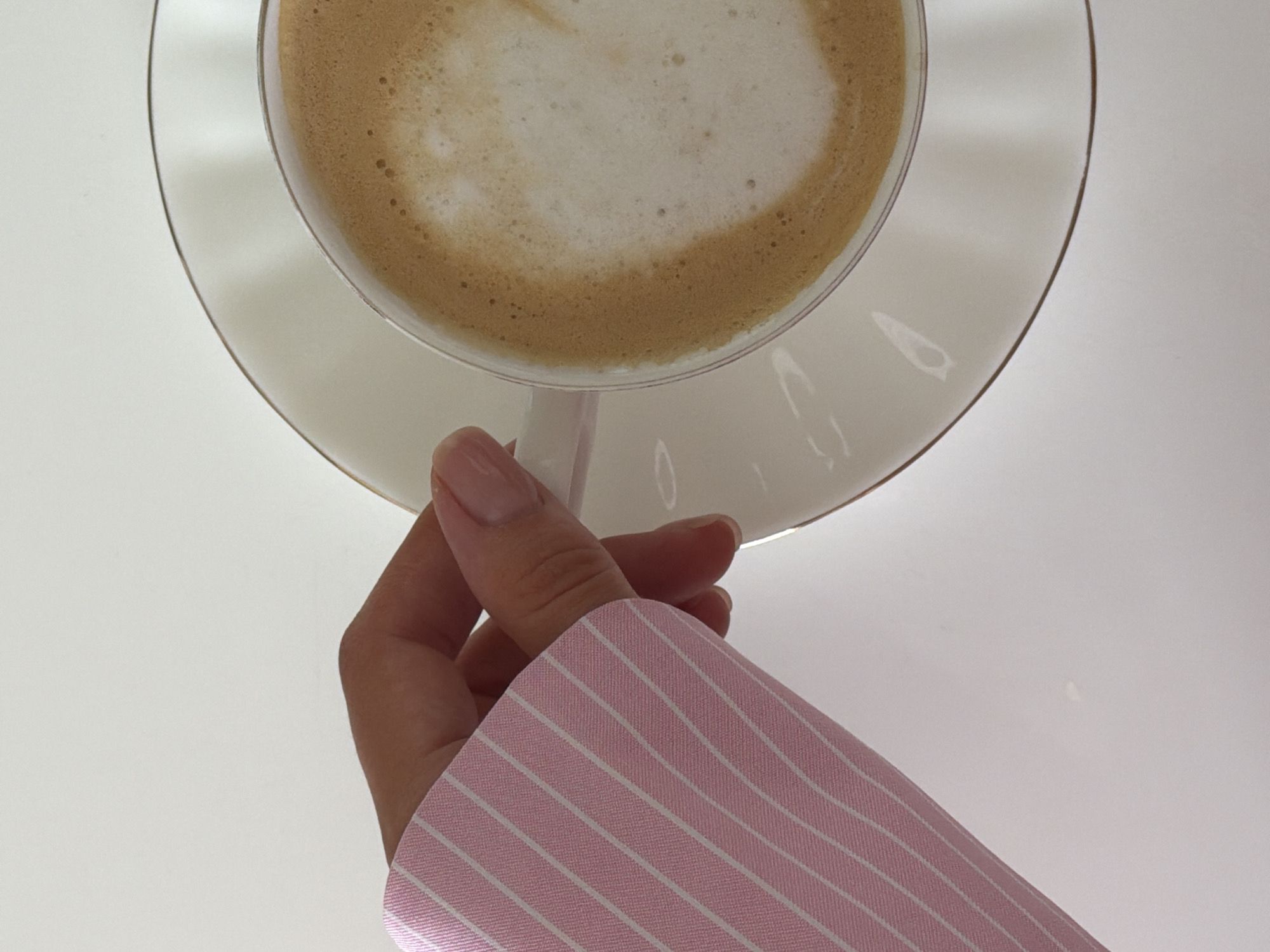 A person wearing a pink and white striped shirt is holding a glass cup of coffee with a saucer. The coffee appears to be a latte or cappuccino with a foamy top. The background is a plain white surface. The location is not discernible from the image, therefore I cannot infer that it is in the city of Khimki, Russia.