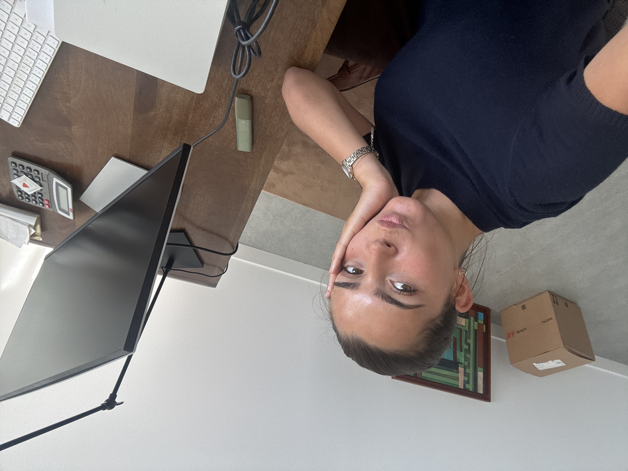 A woman with dark hair pulled back, wearing a dark blue short-sleeved top and a silver watch, is seated at a desk. She is looking directly at the camera with her hand resting on her face. The desk holds a white keyboard, a white Apple laptop, a gray calculator, a white mouse, a green highlighter, and a large computer monitor on a stand. In the background, there's a framed abstract artwork and a cardboard box with "H&M" printed on it. The location is not discernible from the image. It is not possible to determine if the location is in the city of Torres Vedras, Portugal.