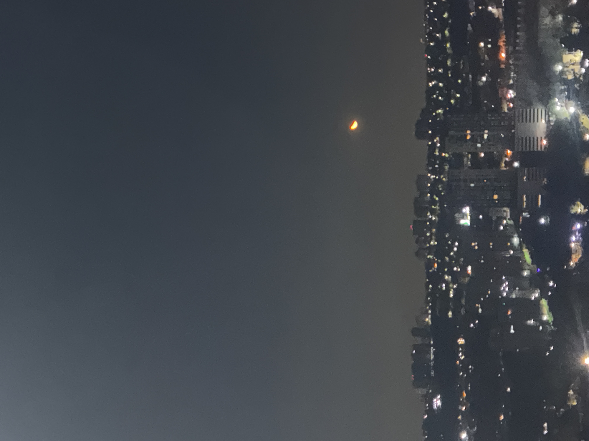 Here's a concise description of the image:
A nighttime shot shows a cityscape on the right, with the dark sky dominating the left. A crescent moon is visible in the sky. The city's lights illuminate buildings and streets. The photograph is taken from a high vantage point, likely from a building or elevated area. The location may be Huixquilucan de Degollado, Mexico.