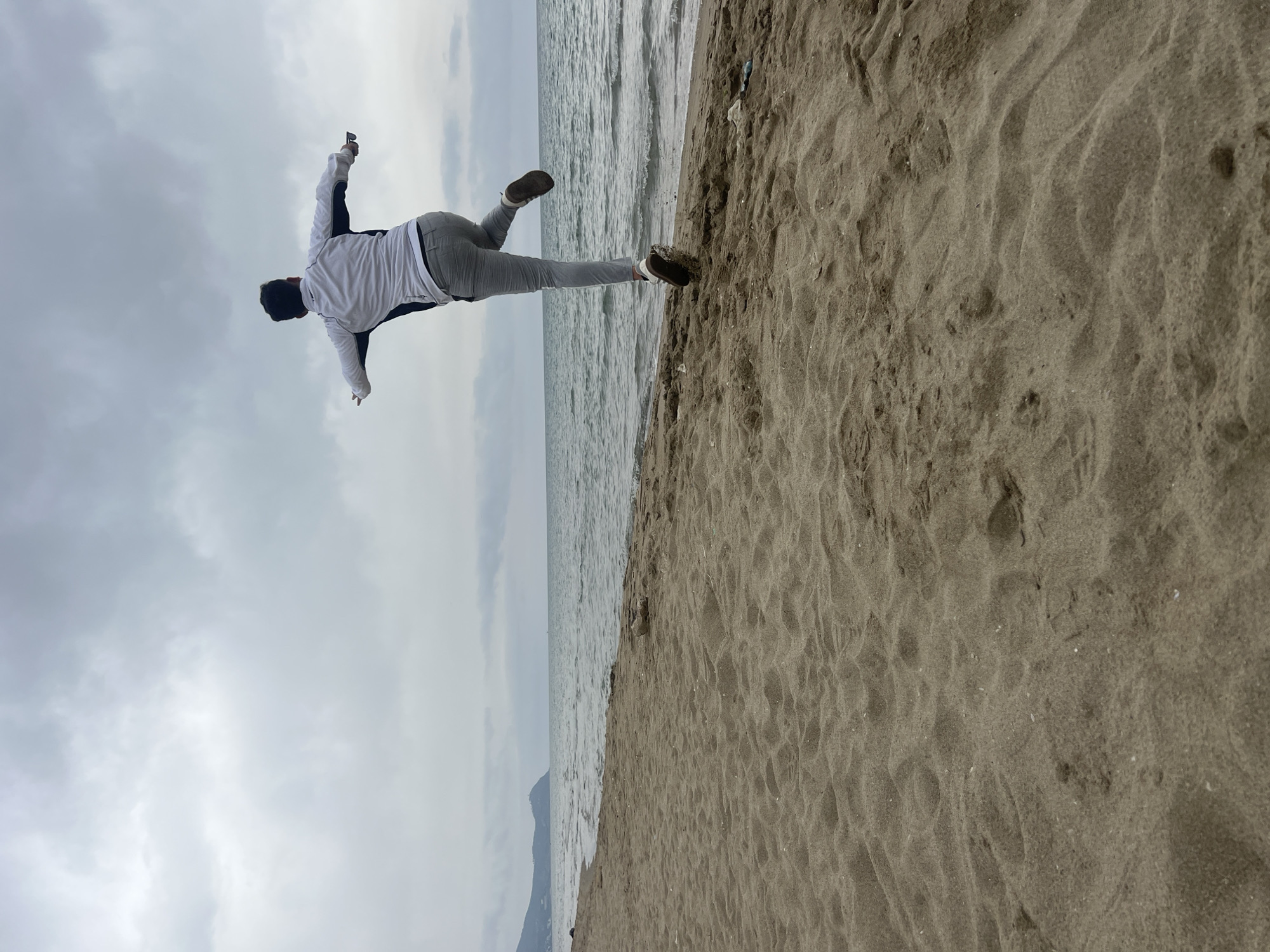 The image captures a person on a beach in Martil, Morocco, frozen in a dynamic, active pose. Viewed from behind, the individual wears a white long-sleeved top with black sections, grey pants, and dark shoes. Their arms are extended outwards, and one leg is bent and lifted backward, creating an impression of balancing, leaping, or mimicking flight.
The scene unfolds under an overcast sky, which appears uniformly grey and cloudy. To the right, a significant portion of the frame is occupied by the sandy beach, exhibiting natural ripples and indentations. To the left, the calm, grey-toned sea meets the shore with gentle waves. A faint, dark silhouette, possibly a distant mountain or landmass, can be seen on the horizon line across the water. The overall color palette is muted, dominated by shades of grey from the sky and sea, and the natural brown of the sand, with the person's attire complementing these tones. No text is visible in the image.