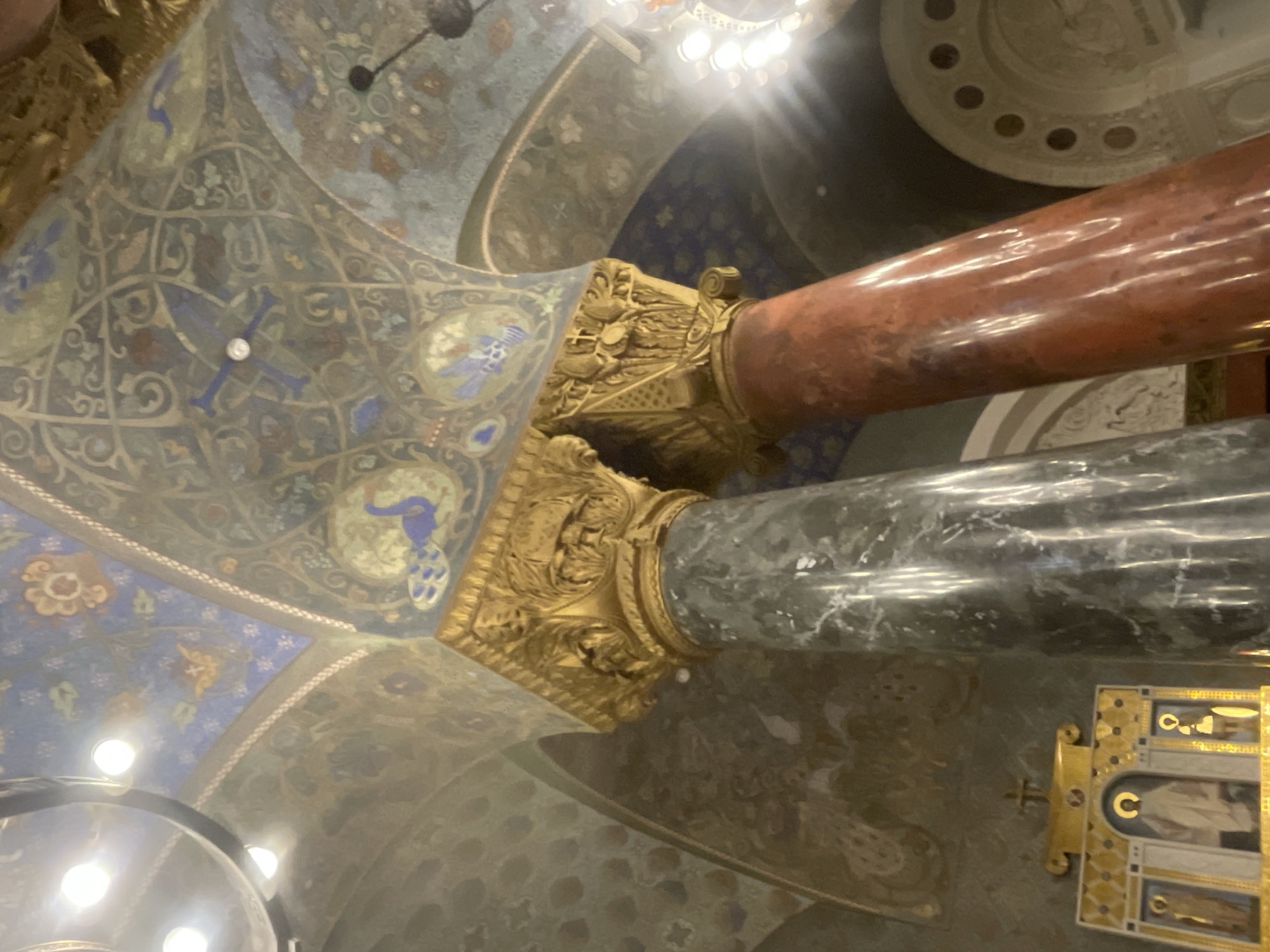 This image shows the interior of the St. Nicholas Naval Cathedral in Kronstadt, Russia. The view is from below, looking up at the ornately decorated domed ceiling and supporting columns. Two large, polished marble columns are prominent in the foreground: one appears to be made of dark green or gray marble, and the other of reddish-brown marble. Both columns are topped with intricately carved golden capitals featuring acanthus leaves and other decorative motifs.
The ceiling is a complex mosaic or fresco, painted in shades of blue, gold, green, and red, depicting various figures, geometric patterns, and what appear to be stylized birds or mythical creatures. The overall impression is one of grandeur and religious artistry.
In the lower left quadrant of the image, a portion of a gilded iconostasis or religious artwork is visible, featuring depictions of saints or religious figures in gilded frames. There are no people visible in the image, and the scene appears to be static and posed, focusing on the architectural and artistic details of the cathedral's interior. The lighting suggests it might be indoors, with some artificial lights reflecting off the polished surfaces and contributing to the overall ambiance.