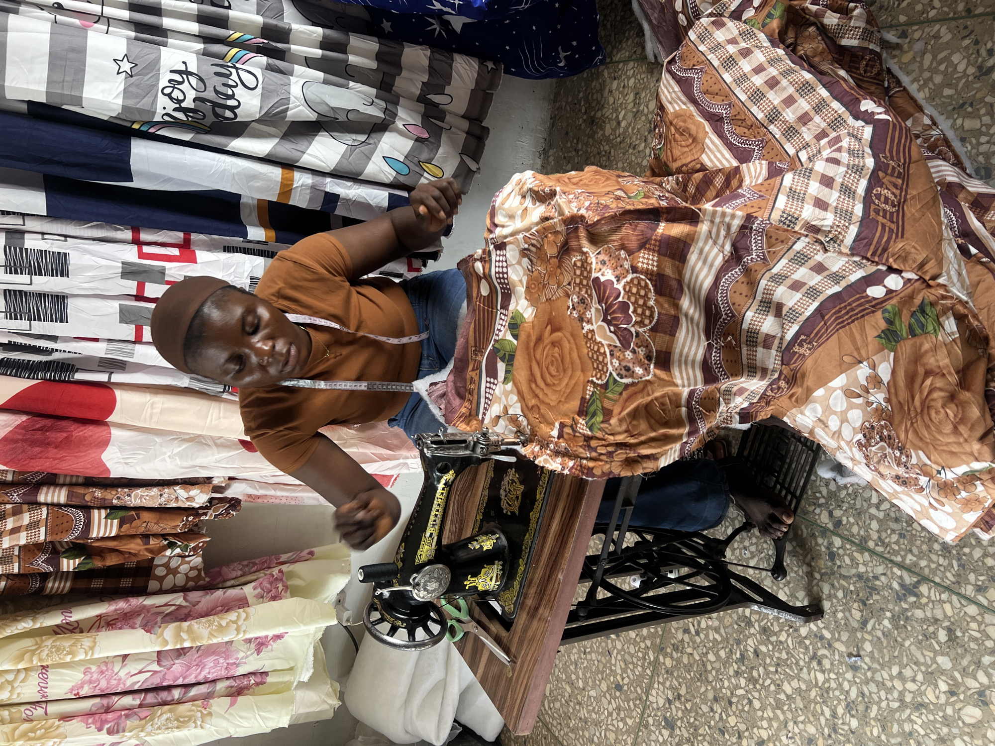 A person is operating a sewing machine in a room in Jos, Nigeria. The person, wearing a brown shirt and blue jeans, is seated and focused on the sewing machine. The machine is vintage and black with gold accents. A colorful, patterned fabric is being sewn, and several other fabrics hang in the background. The room's floor is tiled, and the walls are bare. The lighting appears to be natural, suggesting the presence of a window or open door.