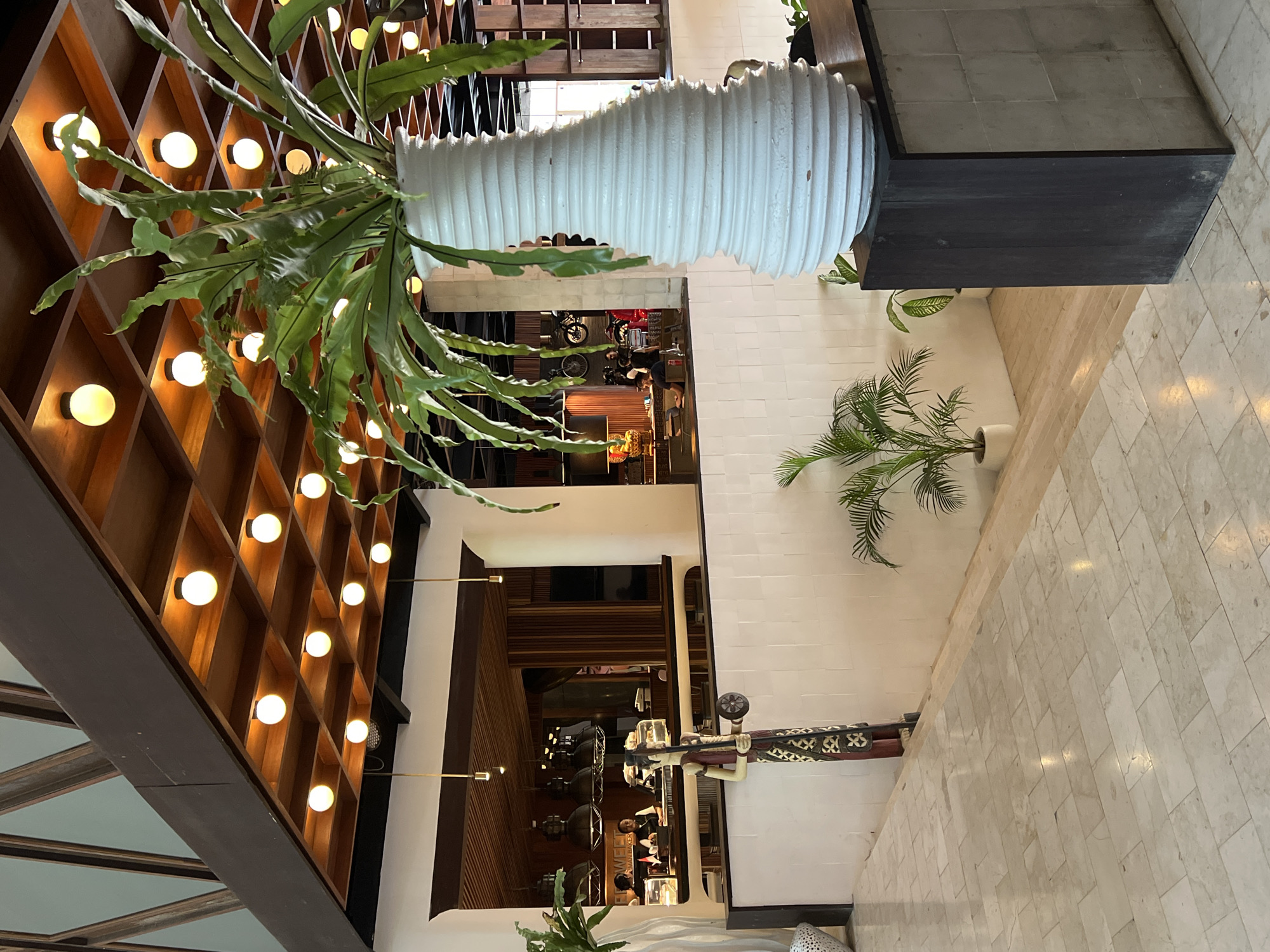 The photograph captures the interior of what appears to be a commercial space, potentially a hotel lobby or upscale restaurant, located in Petulu, Indonesia. The vantage point is from above, looking down into the space.
The scene includes a blend of modern design elements and local Indonesian cultural artifacts. A grid-like structure containing multiple round light fixtures is on the left. A large, white, corrugated vase or planter sits atop a dark, rectangular base, with greenery cascading from it. A smaller palm tree in a white pot is nearby.
In the lower center of the frame, a statue of a figure holding a tray is visible. Below, a glimpse into another area shows people seated at tables, suggesting a dining or waiting area. The flooring is composed of light-colored, rectangular tiles. The walls are primarily white.
The overall lighting is warm, casting light on the wooden elements and illuminating the space. The scene is unoccupied, suggesting it was taken during a quiet moment or before/after operating hours.