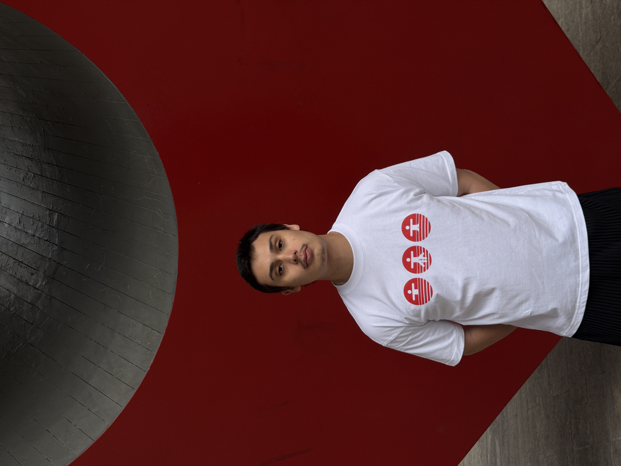 A young man stands against a backdrop of geometric shapes. He is facing the camera, his hands behind his back. The man is wearing a white t-shirt with three red circular logos. The background is composed of a large red surface meeting a curved gray shape on the left, and a light gray area on the lower right. The image has a slightly off-kilter perspective. The location is in an urban area, most likely in New York, USA.
