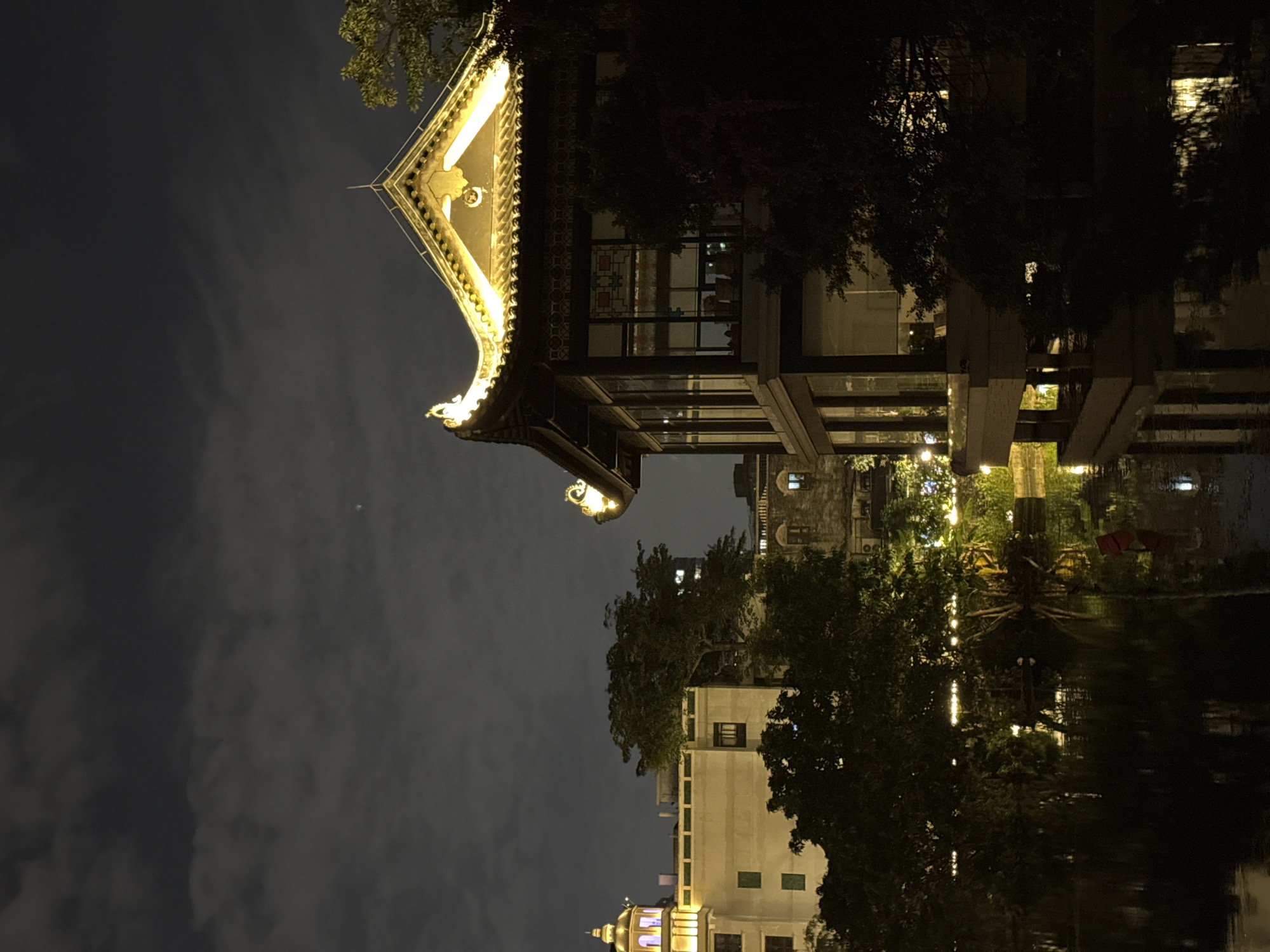 The image depicts a nighttime scene in Liwan District, China. A traditional Chinese building with an ornate, illuminated roof is prominent. The building is reflected in a body of water, likely a lake or canal, creating a mirrored effect. Modern buildings and trees can be seen in the background, also reflected in the water. The sky is overcast and dark.