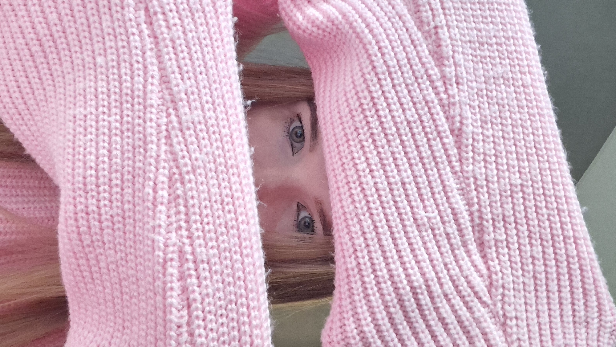 The image shows a close-up view of a person's face, specifically their eyes and part of their forehead and nose, peering through a gap in a pink knitted sweater. The sweater takes up most of the frame. The background is slightly out of focus, showing a hint of light-colored hair. The image is cropped, focusing on the eyes looking directly at the viewer. There is no visible information indicating the location as Chaikovsky, Russia.