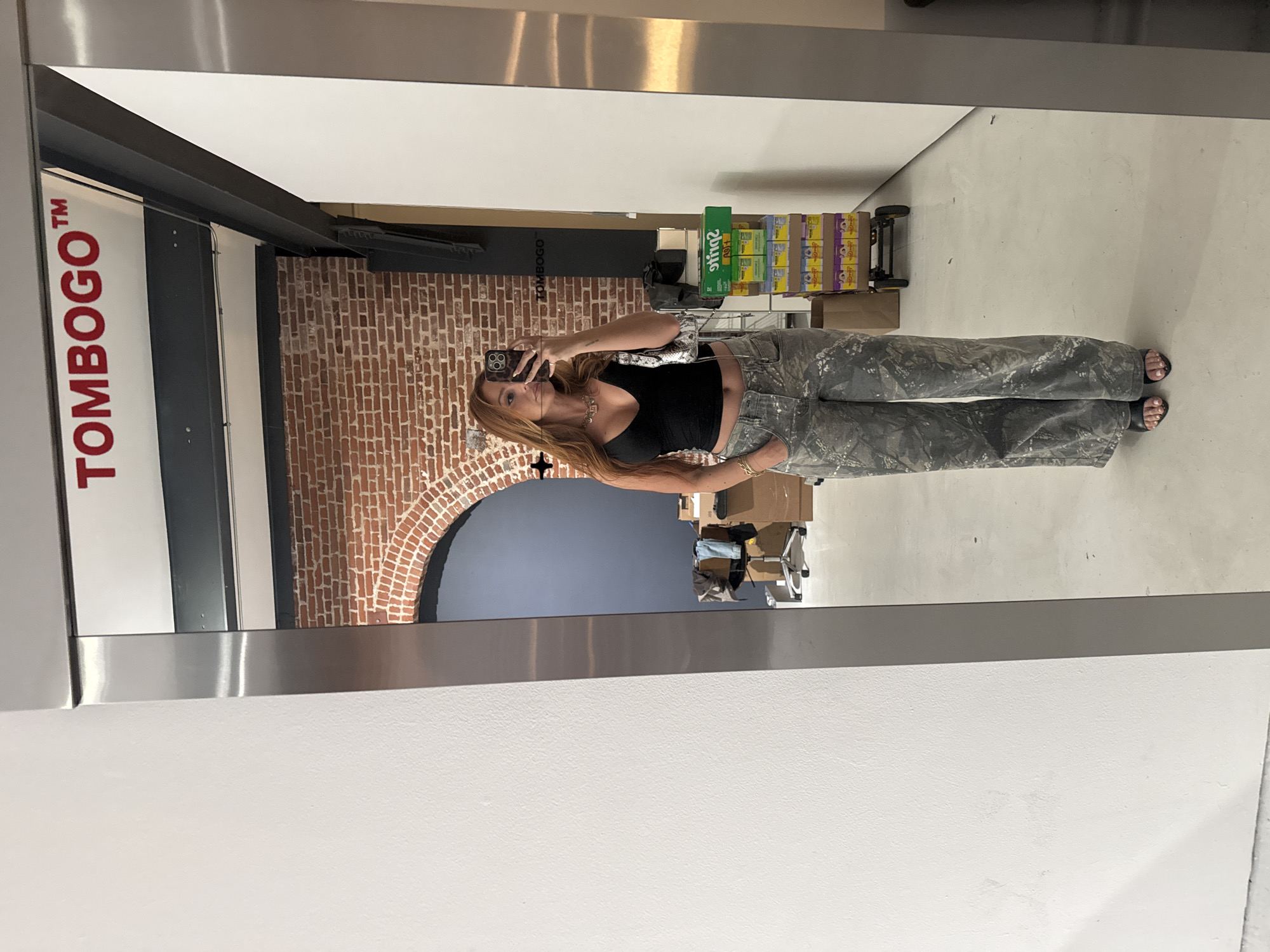 A woman is captured in a full-body mirror selfie within what appears to be an indoor commercial or industrial space, possibly a retail store or warehouse. She stands with her left arm casually bent at the hip and holds a smartphone with her right hand to take the picture, concealing her face. She has long, reddish-brown hair and is wearing a black cropped tank top, wide-leg or straight-leg pants with a greyish camouflage or paint-splatter pattern, and black sandals. Accessories include a gold-colored chain necklace, a bracelet on her left wrist, and a small, light-colored patterned shoulder bag with a dark strap across her body. Her phone case has a leopard print or similar pattern.
The background reflected in the mirror features diverse architectural elements. To the left, a white vertical panel prominently displays the red text "TOMBOGO™". Adjacent to this, there is an exposed red brick wall with a distinct arched opening, indicating a doorway or passage. The space also includes modern white walls, dark metal structural beams, and a light-colored concrete floor. In the further background, stacks of merchandise boxes are visible, with the green packaging of "Sprite" clearly identifiable on some. A black hand truck is also present near the boxes. The lighting appears bright and even, suggesting a well-lit interior environment during daytime.