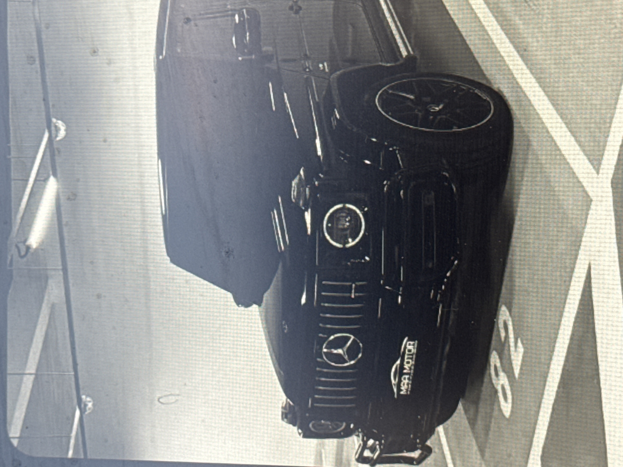 A black Mercedes-Benz G-Wagen is parked in what appears to be an indoor or underground parking garage in Varna, Bulgaria. The image is rotated 90 degrees clockwise, showing the front of the vehicle predominantly. The car is sleek and black, featuring the distinct Mercedes-Benz logo prominently on its grille, flanked by round headlights. Below the grille, a badge or custom license plate holder reads "MAA MOTOR." The floor of the parking space is marked with white lines and the number "82," indicating a specific parking spot. The overall lighting suggests an artificial indoor environment. The image quality is somewhat grainy, possibly indicating it's a photo taken of a screen. No people are visible, and the car appears to be stationary.