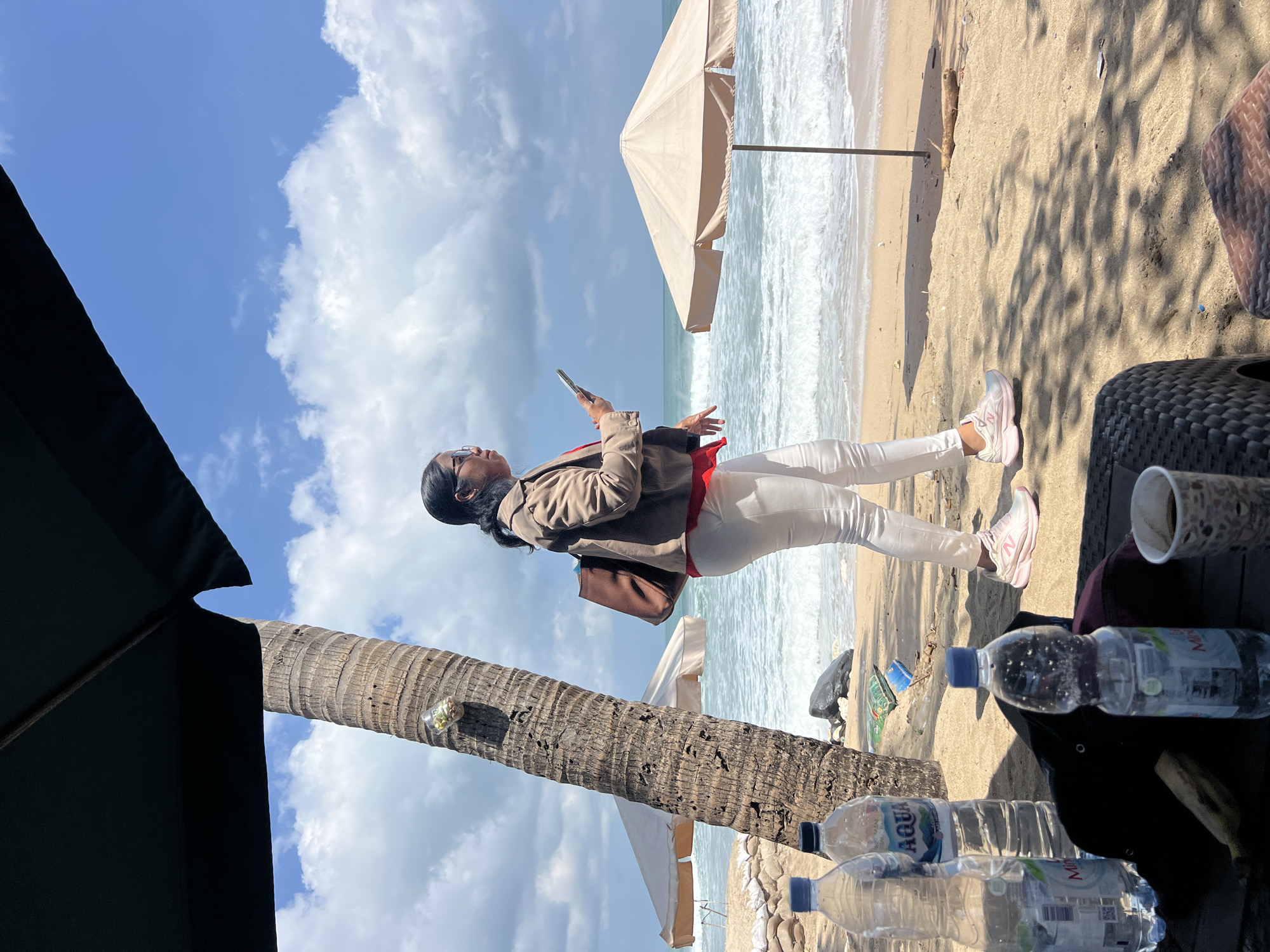 A woman in a tan jacket, red shirt, and white pants stands on a sandy beach in Kuta, Indonesia, holding a mobile phone. She is positioned between a palm tree and a light-colored beach umbrella. The ocean with waves is visible behind her, and the sky is blue with scattered white clouds. In the foreground, there are two plastic water bottles and a dark bag on a woven surface, with a cup of coffee to the right. Several sandbags are visible near the palm tree.