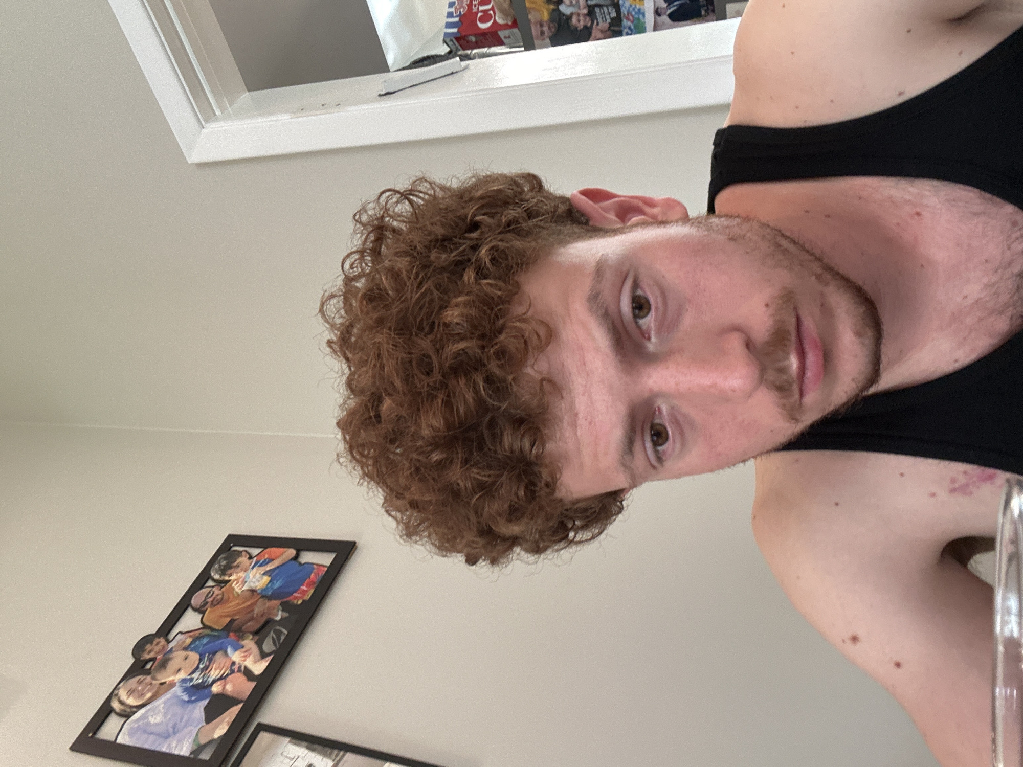 A medium, slightly rotated close-up shot features a man with curly brown hair, light skin, and a light brown beard. He is wearing a black tank top. The background is a plain, light-colored wall. A picture frame with a collage of people is visible in the lower left corner of the frame. At the top of the frame, a window can be seen with miscellaneous objects on the window sill. The location is suggested to be in Pepper Pike, United States of America.