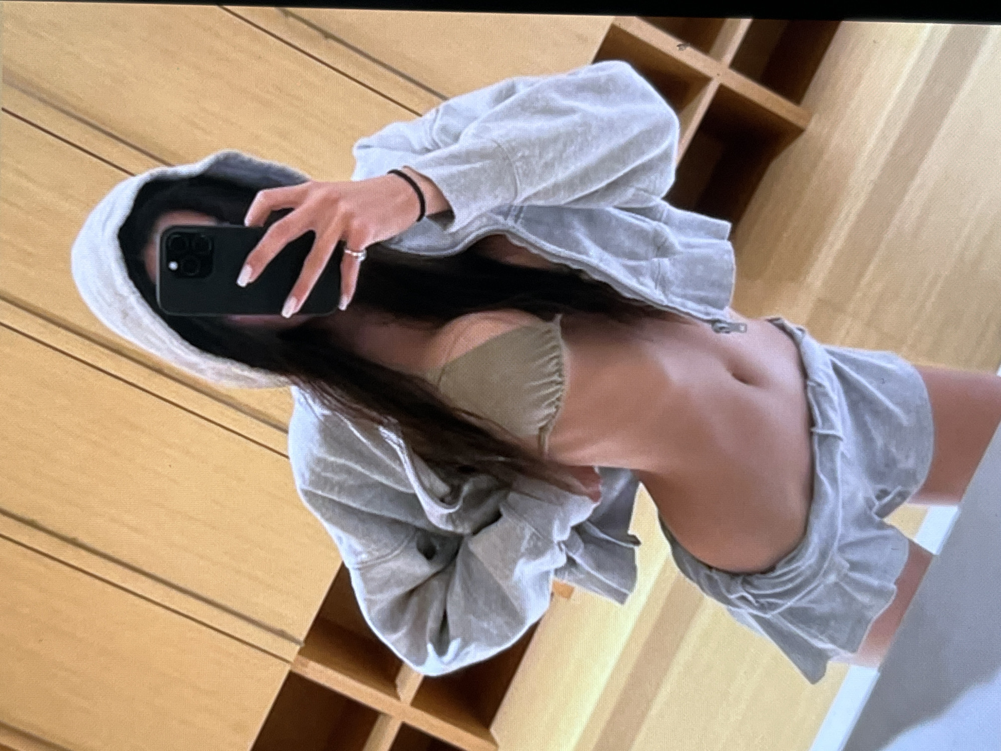 A young woman is taking a selfie in what appears to be a closet or dressing room. She is wearing a grey zip-up hoodie, a light brown bikini top, and ruffled grey shorts. Her long, dark hair is down. She is holding a black smartphone in her right hand, which is reflected in a mirror, capturing her image. Her left hand is pulling at the hoodie. The background consists of light-colored wooden cabinetry with visible shelving. The lighting is somewhat dim.