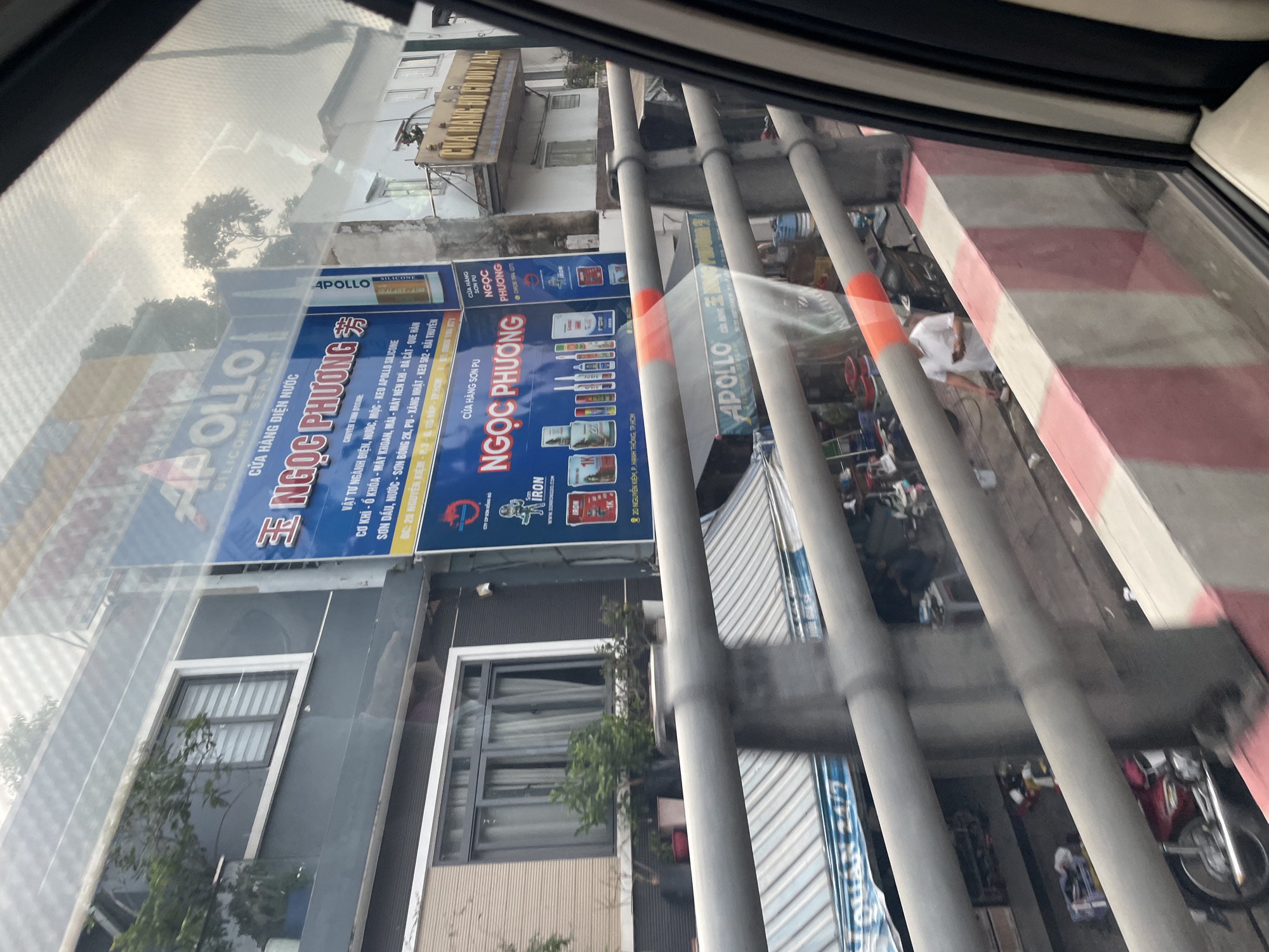 The image shows a street scene in Thu Duc City, Vietnam, viewed from inside a vehicle. A large blue billboard advertising "NGỌC PHƯƠNG" and "APOLLO SILICONE SEALANT" is prominently displayed on the side of a building. The billboard lists various products and services related to construction and sealants. Below this billboard, another sign reads "CỬA HÀNG SƠN PU NGỌC PHƯƠNG". Further down the street, a smaller yellow sign can be seen on another building that appears to be a shop. A red motorcycle is parked on the street. The scene also includes a concrete barrier with red and white stripes, which is likely part of an overpass or elevated road. A person in a white shirt is visible standing on the street below the overpass. The sky is overcast, indicating cloudy weather. The overall impression is a typical urban street view with commercial signage and everyday street life.