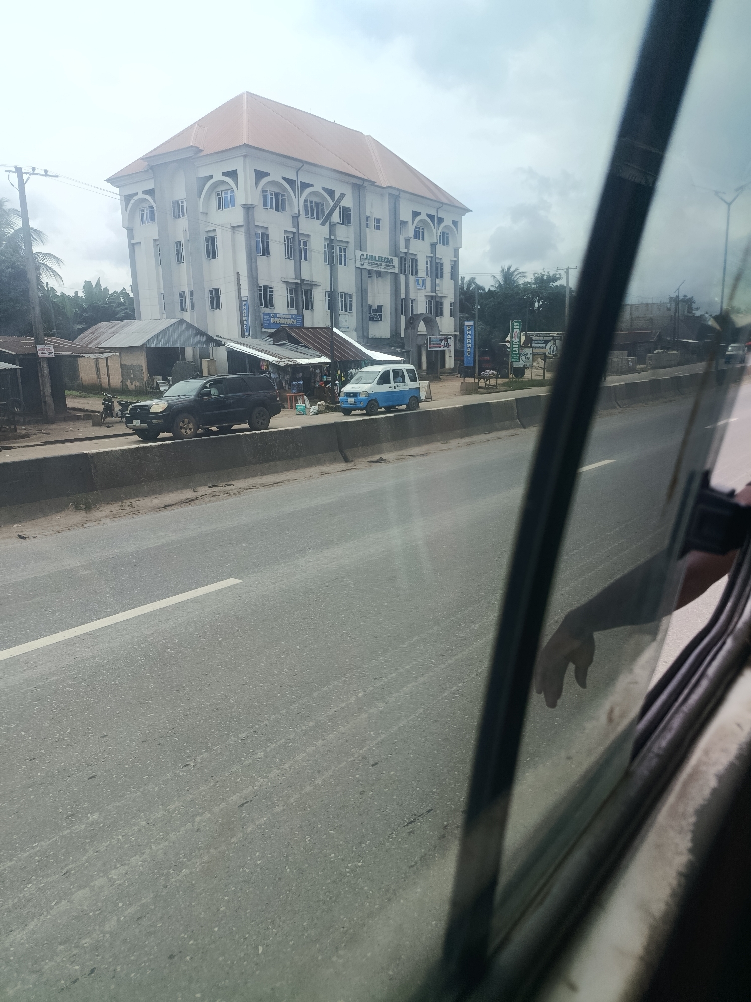 A medium shot, captured from a vehicle's interior, shows a four-story building with a distinctive arched facade on the left side of a road in Orogwe, Nigeria. The building is pale beige with a reddish-brown roof and features numerous windows. A sign on the building reads "JUBILEE CARDIAC AND SPECIALIST HOSPITAL" and "PHARMACY." In front of the building, several smaller structures with corrugated metal roofs are visible, some appearing to be shops or market stalls. A black SUV is parked on the side of the road in front of these structures. Further down the road, a white and blue minivan is visible. The road in the foreground is asphalt with a white lane marking. Trees and a cloudy sky are present in the background. The right side of the image is dominated by the reflection of a hand and arm in the car window, obscuring the view of the road and surroundings.