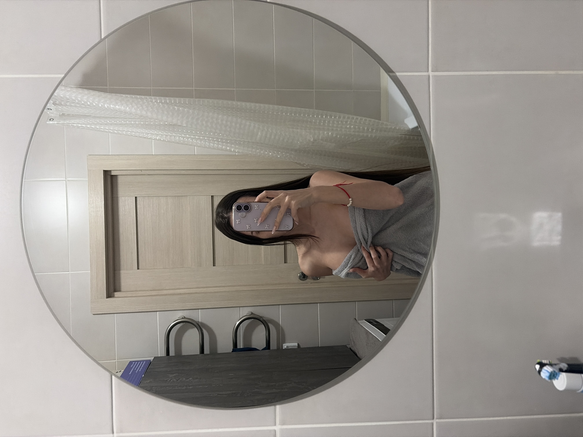 A person is depicted taking a mirror selfie in what appears to be a bathroom. The subject, likely a young female, is seen from the chest up, wrapped in a light grey towel. Her face is obscured by a smartphone, which she holds with both hands. The phone case is light purple or pink with white floral or butterfly patterns. A red string bracelet, possibly with a small white bead, is visible on her right wrist. Her long dark hair falls over her shoulder and arm.
The setting is a bathroom with light grey or white tiled walls. Reflected in the round mirror are parts of the bathroom: a light wooden door frame, a dark grey counter or vanity with two chrome U-shaped fixtures (possibly towel or toothbrush holders), and a clear, textured shower curtain with a bubble-like pattern. A small blue card or brochure is visible on the counter. Outside the mirror's reflection, on the tiled wall to the right, a toothbrush and a white tube (possibly toothpaste) are partially visible. The lighting suggests an indoor setting, likely a hotel or private residence. No specific location cues for Saint Petersburg, Russia, are visually discernible from the image. No legible text is visible on any objects.