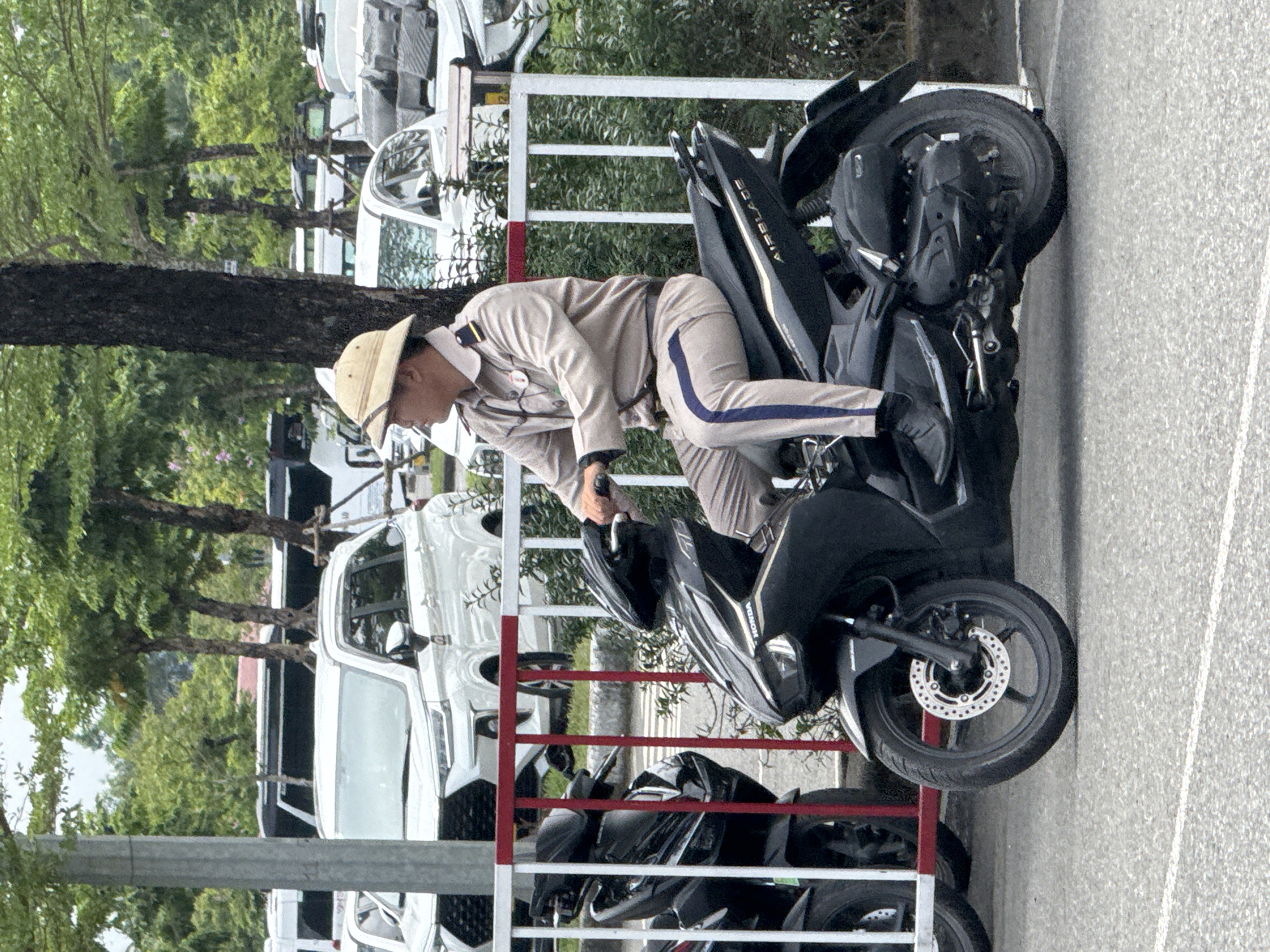 A uniformed official wearing a pith helmet is mounting a black Honda Airblade motorcycle on a city street. The motorcycle is parked against a red and white metal barrier. Behind the barrier, several white vehicles are visible in a parking area adjacent to trees. The location is identified as the city of Đà Nẵng, Vietnam.