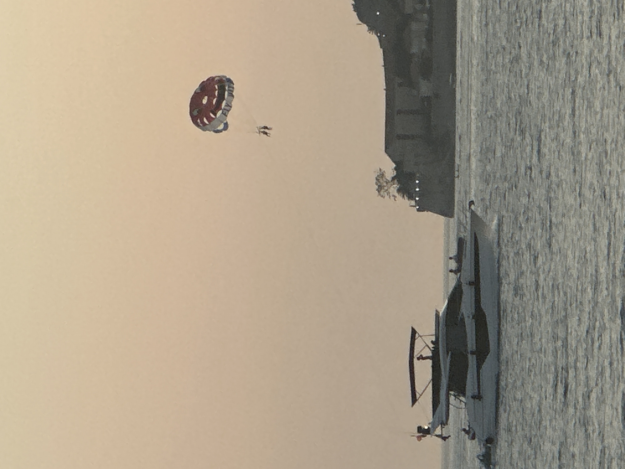 The image captures a parasailing activity in the waters near Budva, Montenegro. In the foreground, a white motor yacht with a dark stripe is moving across the sea, carrying several individuals on its deck. The yacht has a canopy providing shade. Further in the background, two people are visible under a large, red and white parachute, being towed by a boat (not fully visible). A cluster of buildings and a rocky coastline, likely part of Budva's historic old town, are situated on the right side of the image. The sky is a hazy, pale orange, suggesting either dawn or dusk. The water is choppy with visible ripples.