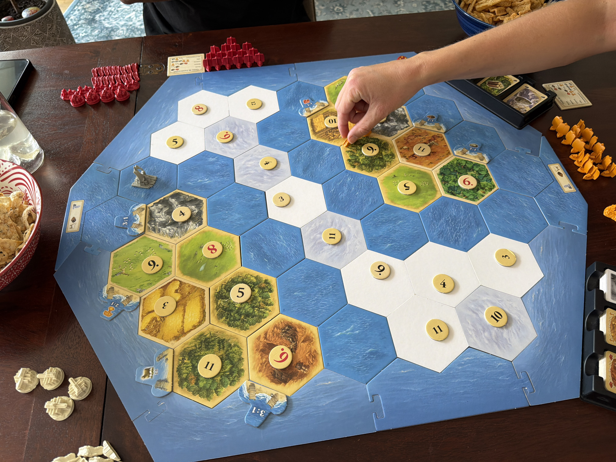This media file captures a close-up view of a game of Settlers of Catan being played on a wooden table. The game board, composed of hexagonal tiles depicting land and sea, is the central focus. The tiles represent various resources like forests, fields, and mountains, indicated by corresponding imagery and numbered tokens.
Several players' hands are visible, interacting with the game. One hand is placing a resource card onto the board, while another hand is about to draw or place a game piece. Scattered around the board are game pieces: red settlements and roads on the left, and orange pieces, likely ships or development cards, on the right. There are also white pieces, possibly also settlements or roads, in the lower left corner. A bowl filled with what appears to be potato chips is visible on the left side of the image, suggesting a casual gathering. In the background, a person wearing a black shirt is partially visible, indicating the presence of multiple players.
The lighting suggests an indoor setting, likely during the daytime, given the ambient light. The wooden table and the general arrangement imply a home or recreational setting. The atmosphere appears relaxed and engaged, typical of a board game session.
Visible text includes numbers on the game tiles (e.g., 11, 9, 5, 6, 8, 10, 3, 4, 12) and trade port designations on specific tiles (3:1, 2:1). A small card with text and icons, likely related to game rules or victory points, is also present near the red game pieces.