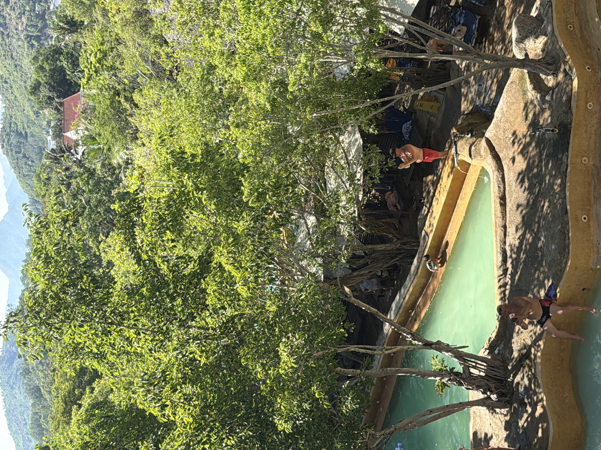 This is an outdoor scene at a resort in Tay Nha Trang Ward, Vietnam, featuring a naturalistic swimming pool area surrounded by lush green trees and foliage. In the foreground, a man in black swim trunks is sitting on the edge of the pool, holding a phone to his ear. To his right, partially obscured by trees and shadows, another man in red shorts stands near the pool's edge, also appearing to be holding a phone. A small child in dark shorts is seen near the pool's edge in the mid-ground, appearing to be stooping or playing. Several lounge chairs and shaded areas are visible in the background, suggesting a relaxed atmosphere. The water in the pool is a clear turquoise color. In the distance, rolling green hills and mountains are visible under a bright sky, with the tops of some buildings peeking through the trees.