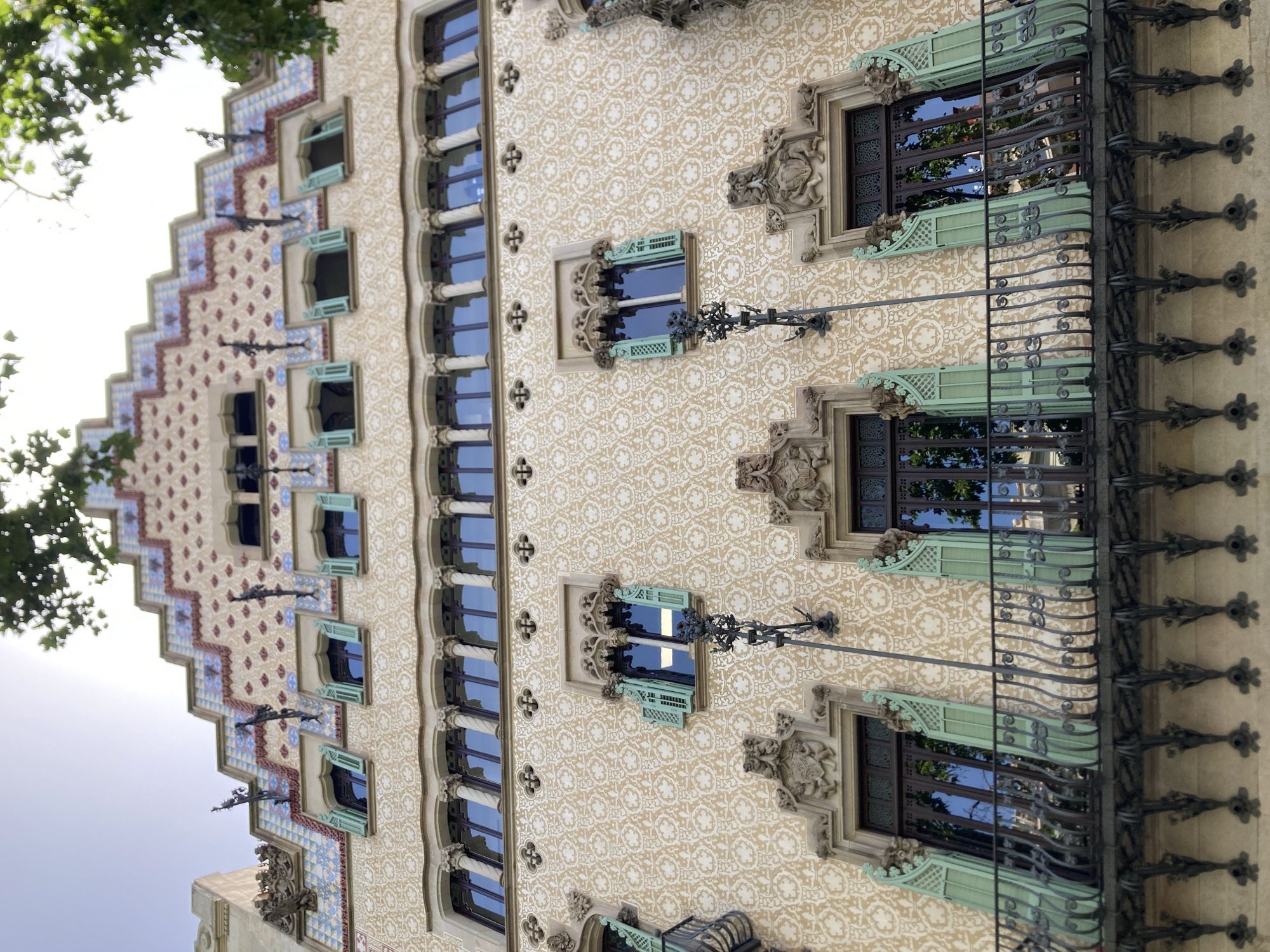 The image captures the facade of Casa Batlló, a renowned building located in Barcelona, Spain, designed by Antoni Gaudí. The building's distinctive organic and undulating architecture is evident, particularly in its upper levels and the characteristic tiled roofline that resembles dragon scales.
The photograph is taken from a low angle, looking up towards the building's facade. The facade is adorned with intricate patterns and decorative elements. Various windows are visible, many with light green shutters, some of which are open. The building's exterior is richly ornamented with sculpted details above some windows and a decorative balcony runs across the lower visible section of the facade. The balcony features a wrought-iron railing with decorative metalwork. The facade itself is covered in a textured pattern of circular motifs, rendered in a light, earthy tone. Above this, different levels of windows are visible, some with columns and decorative stonework. The very top of the building displays a unique roofline with a stepped design, decorated with colorful tiles in geometric patterns. Foliage from trees partially obscures the upper right portion of the building. The sky is visible at the top of the image, appearing bright and possibly overcast. No people are present in the frame.