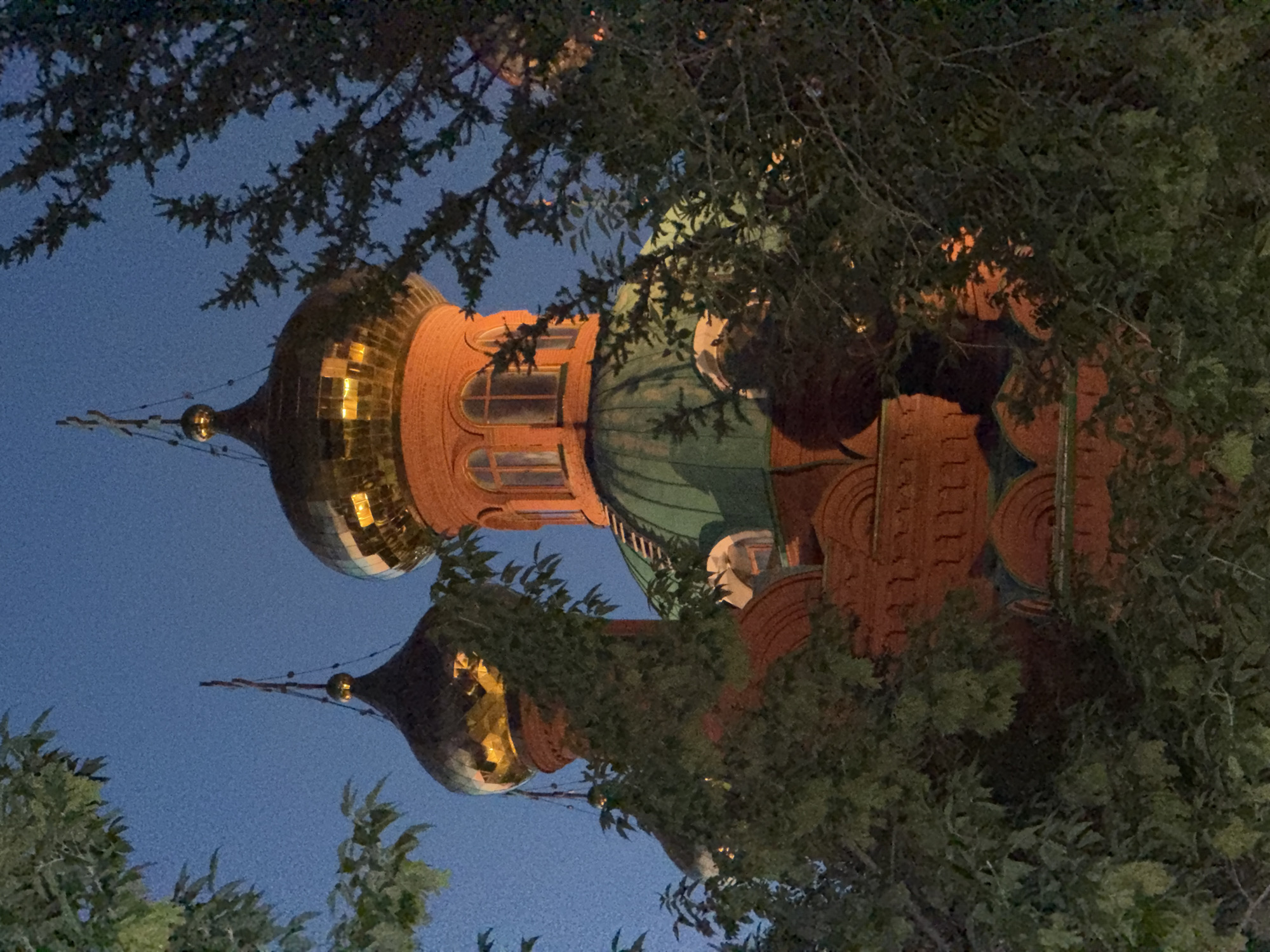 A low-angle, medium shot shows the domes of a red brick church, partially obscured by the foliage of trees. The sky is a deep blue. Two onion domes, one gold and one a muted green, are visible, with the gold dome reflecting warm light, suggesting interior lighting. The gold dome is topped with a cross. The red brick structure has arched windows and decorative brickwork. The image was taken at night or dusk. The church is identified as the Holy Assumption Cathedral in Pavlodar, Kazakhstan. There are no people visible in the image.
