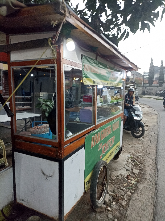 A food cart, with a white exterior and a wooden frame, is parked on the side of a paved road. The cart has a large glass window displaying various food items. On the side of the cart is a green banner with white lettering that reads "AYAM GORENG JAMUR DE JONTOR". A person, wearing a helmet and black clothes, is standing near the cart on a black scooter. The scooter has a black license plate and is parked on the side of the road. There are trees and vegetation in the background. The scene is located in Jatinangor, Indonesia, at the coordinates -6.936 latitude and 107.774 longitude. The cart is situated on the edge of the paved road, facing a direction perpendicular to the road. There is an individual standing on the left side of the cart, partially obscured by the window. The person appears to be interacting with the cart. The cart is situated on the edge of the paved road, facing a direction perpendicular to the road. The environment is quiet and residential, with green vegetation on the left side of the road and a residential street on the right. The scene is brightly lit, indicating that it is likely daytime.