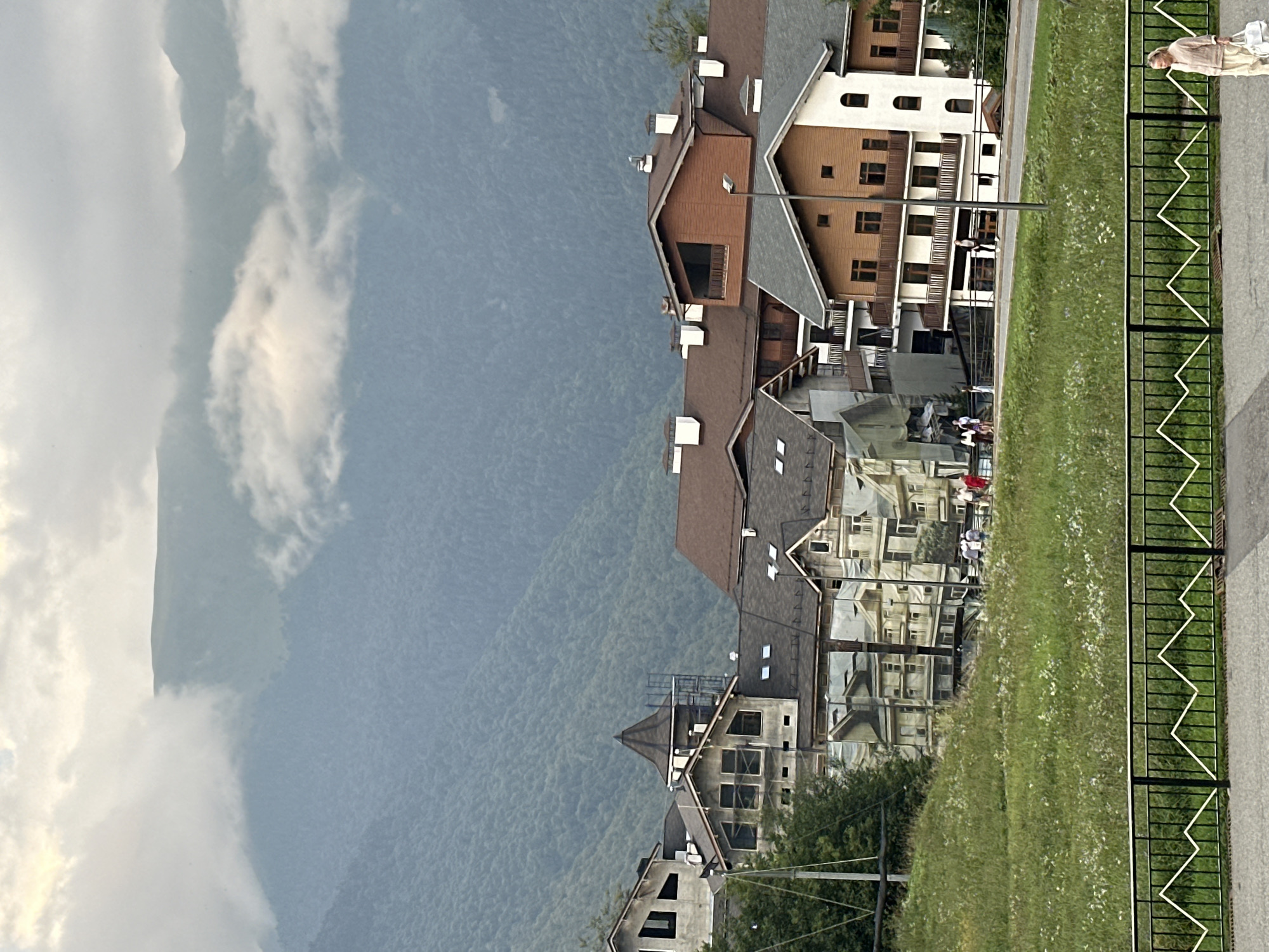 The image depicts a view of residential buildings nestled against a backdrop of lush green mountains partially obscured by clouds. The buildings appear to be of modern architecture, with varied rooflines and materials, including wood and stucco. Some structures are under construction, indicated by scaffolding and protective coverings. A paved pathway runs in the foreground, bordered by a black metal fence with a decorative mountain-like pattern. A woman, dressed in light-colored clothing and carrying a bag, is walking on the pathway. In the middle ground, several people are visible walking along a road that winds between the buildings. The overall setting suggests a resort or mountainous residential area, consistent with the location of Estosadok, Russia. The sky is overcast, contributing to a diffused light over the scene.