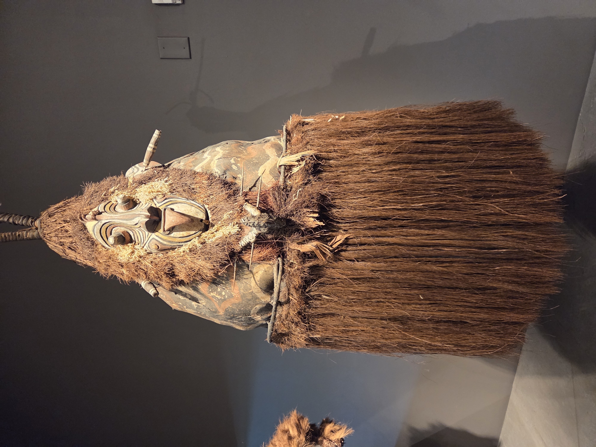 An elaborate tribal mask, likely made of wood, straw, and natural fibers, is displayed against a dark gray wall in a museum setting in Santa Ana, California. The mask features a prominent face with stylized, concentric circular eyes, a wide mouth with visible teeth, and a prominent nose. It is adorned with brown, fibrous material around the head and shoulders, resembling hair or straw. A long, skirt-like fringe of dark brown fibers hangs from the lower portion of the mask, reaching almost to the floor. The mask is suspended from above, with visible darker elements at the top suggesting it is part of a larger costume or effigy. Two thin, light-colored rods protrude from the sides of the mask. The environment is dimly lit, with a shadow cast by the mask on the wall behind it. Another smaller, similarly adorned object is partially visible to the left of the main mask.