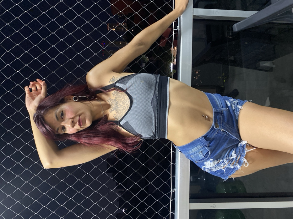 A woman with reddish-brown hair leans against a fence and window frame. She wears a gray sports bra and ripped denim shorts. A tattoo adorns her chest. The background features a dark night sky.