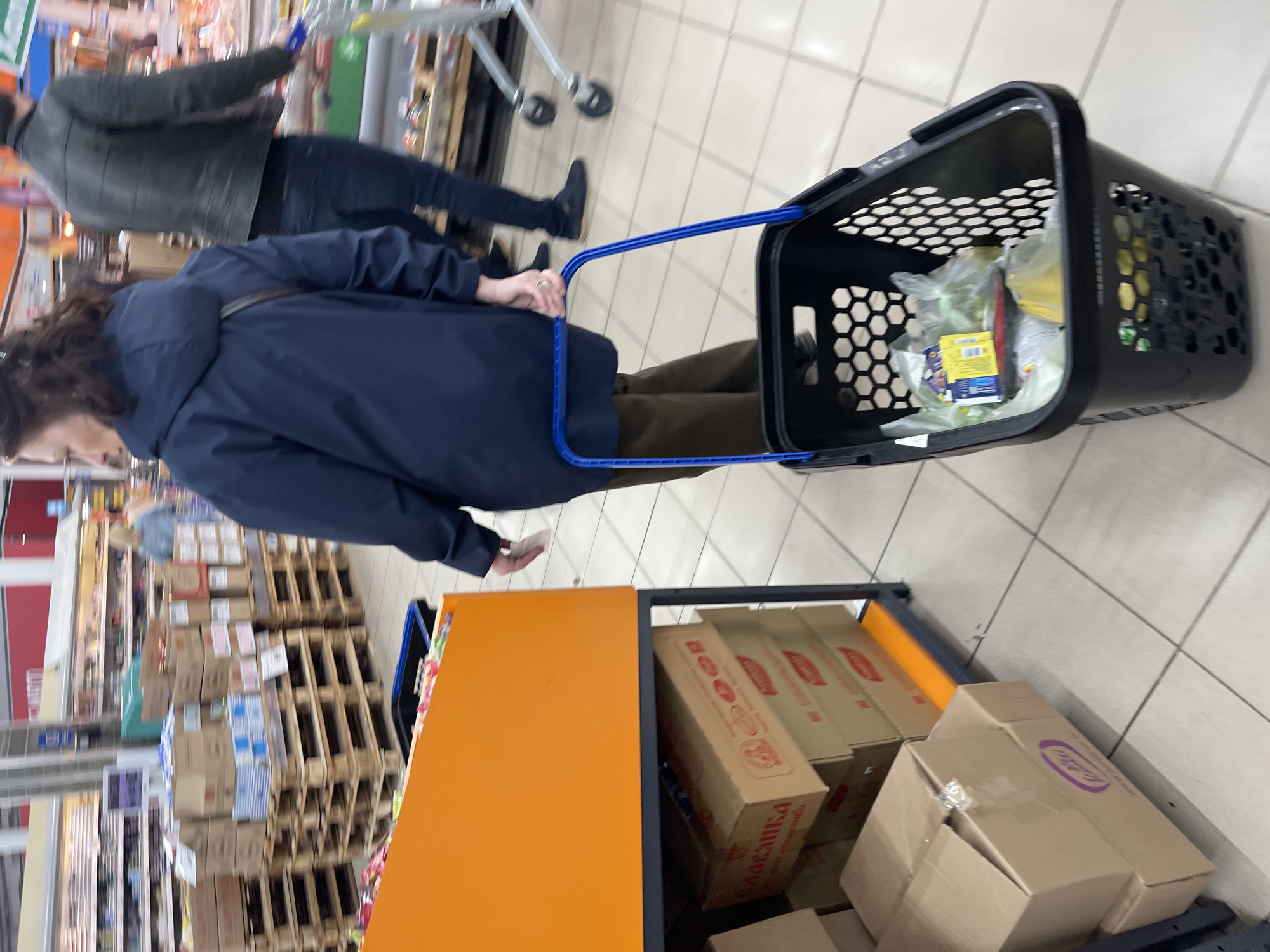 In a retail setting in the city of Shcherbinka, Russia, a customer wearing a blue jacket pulls a black plastic shopping basket with a blue handle containing a selection of grocery items. Another customer in a gray jacket and dark pants walks nearby. A shelf with stacks of packaged goods is adjacent to the customer with the basket. The background is filled with more aisles and products typical of a grocery store.