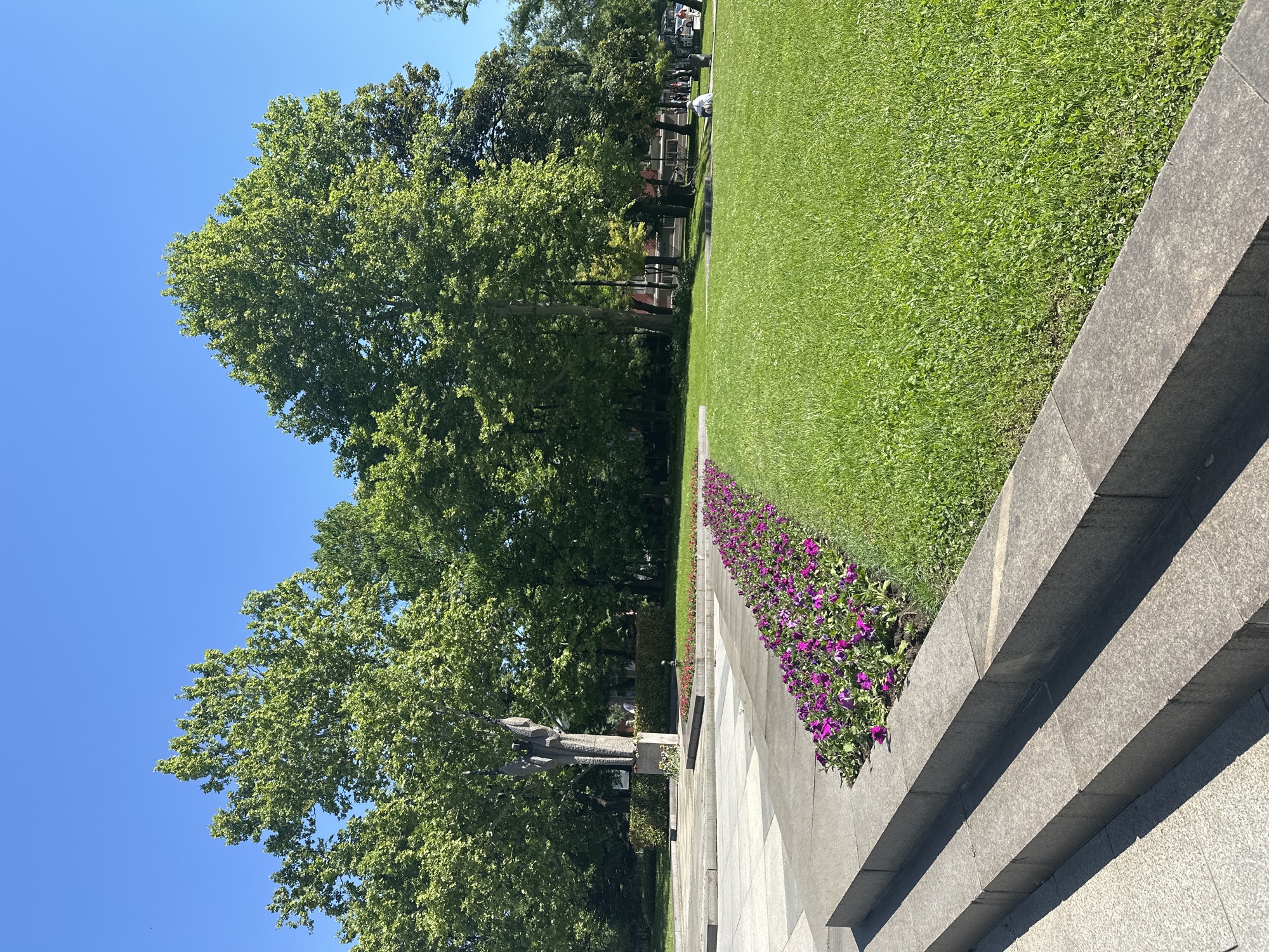 The image shows a section of the Alley of Cosmonauts in Sofia, Bulgaria. A stone monument is partially visible to the left, with large trees providing a backdrop. A well-manicured grassy area extends to the right, bordered by a stone ledge planted with purple flowers. A multi-tiered stone structure is in the foreground. The sky is clear and blue. People are visible in the background, near the treeline.
