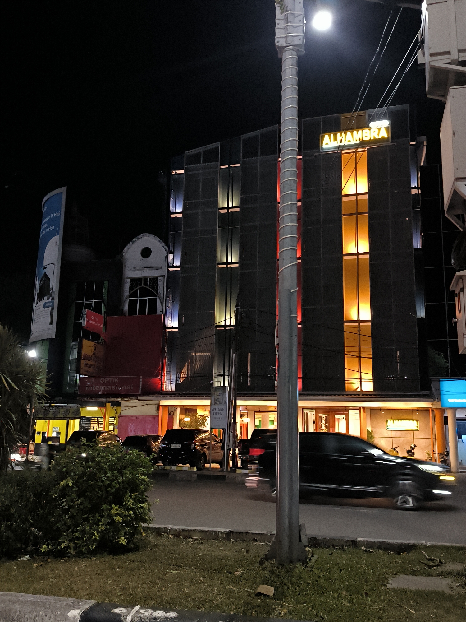 This is a night-time, medium-shot, eye-level photograph taken in front of the "ALHAMBRA" building in Banda Aceh, Indonesia. The building, a modern structure with a dark facade featuring vertical lighting elements in shades of orange and white, is illuminated by its own lights and the streetlights. A prominent yellow neon sign reading "ALHAMBRA" is visible on the upper portion of the building. In the foreground, a tall, utilitarian lamppost with a light fixture at its top stands to the left, partially obscuring the view of the building. The street in front of the building is paved, and a sidewalk area with a low concrete barrier separates it from a grassy area with some low bushes. Several dark-colored vehicles are visible on the street, with one black SUV in motion in the right half of the frame, its headlights blurred by motion. Other vehicles appear to be parked. A large vertical billboard is partially visible on the left side of the image, advertising something in Indonesian. Signs for "OPTİK Internasional" and other businesses are mounted on the lower part of the building. The overall atmosphere is that of a typical urban street scene at night, with artificial lighting illuminating the commercial establishments.