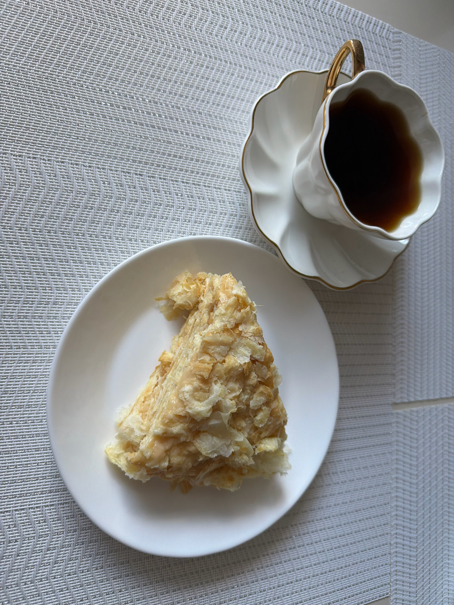 A slice of layered cake, possibly Napoleon cake, sits on a white plate. The cake is light in color with visible flaky layers and cream filling. To the right of the plate is a white teacup with a gold rim, filled with a dark liquid, likely tea or coffee, resting on a matching saucer with a gold scalloped edge. The setting is on a textured white placemat with a woven pattern, possibly on a table. The overall scene suggests a simple, elegant presentation of dessert and a beverage. The image does not provide information to determine its location in Koszalin, Poland.