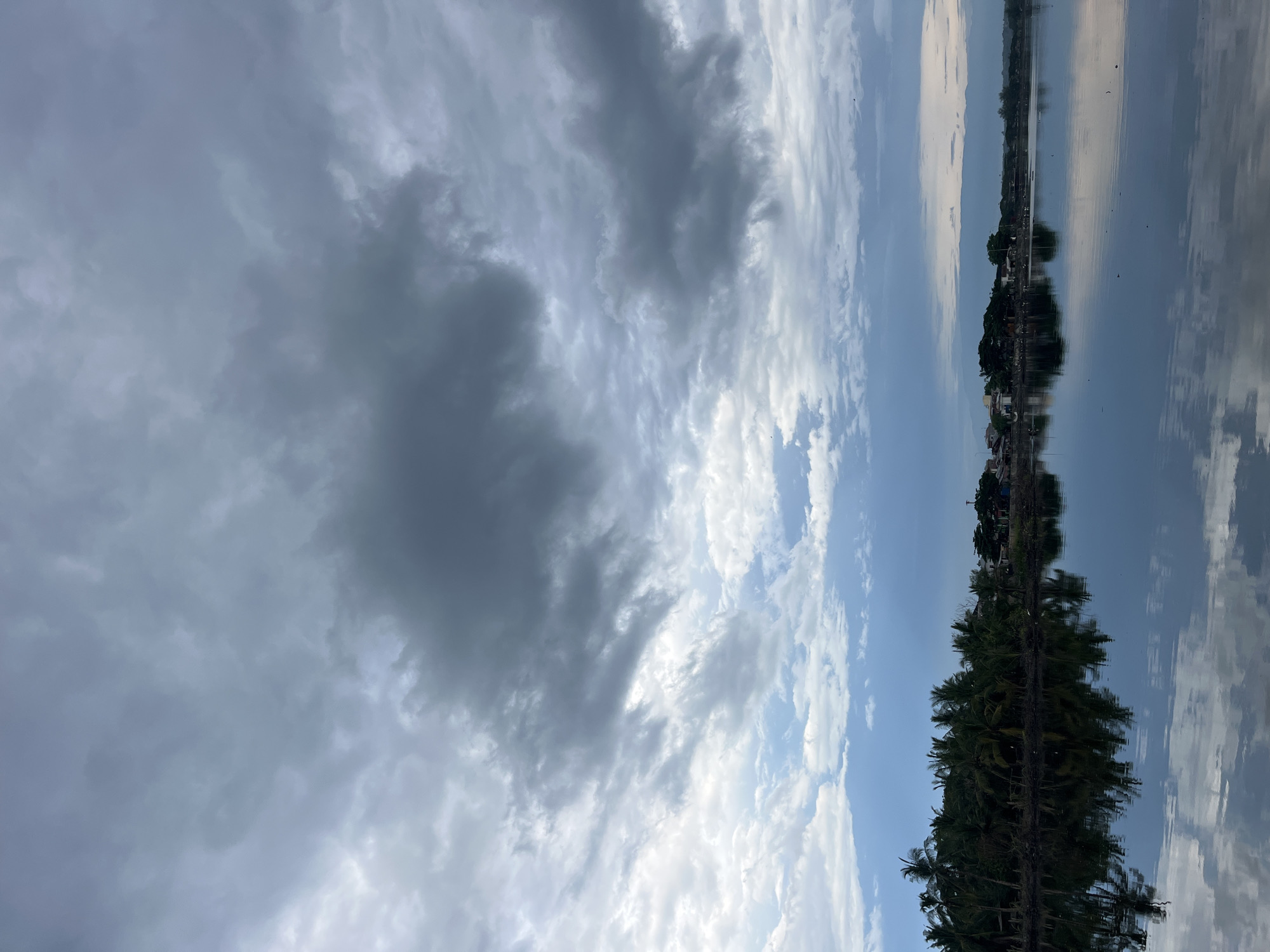 The image shows a water scene near Lhokseumawe, Indonesia. The foreground is dominated by a body of water reflecting the sky and trees. The sky is filled with clouds, ranging from dark gray to bright white. The shoreline is visible in the background, with vegetation including palm trees and what appears to be a row of houses or buildings. The clouds are also reflected in the water.