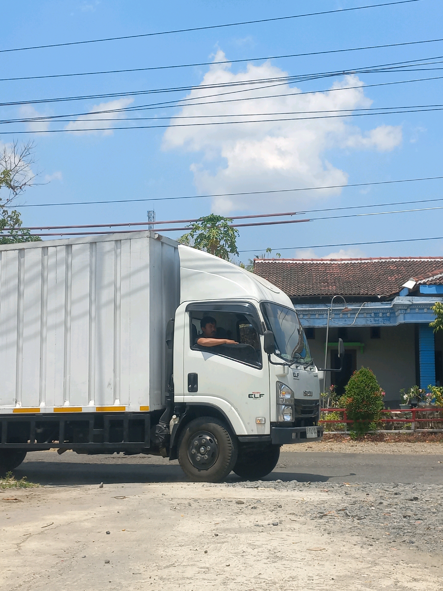 The image captures a side view of a white Isuzu Elf truck driving on a paved road in Baturetno, Indonesia. The truck's cargo box is predominantly white with vertical paneling. The driver, a male with dark hair and a mustache, is visible through the driver's side window, resting his right arm on the window sill. The truck has a black chassis and wheels, with yellow reflective tape along the lower side of the cargo area. In the background, a house with a red tiled roof and blue trim is visible, along with a small, green conical bush and some red fencing. The sky is bright blue with scattered white clouds and overhead power lines traverse the upper portion of the frame. The foreground consists of a dusty, unpaved surface littered with small stones and debris.
